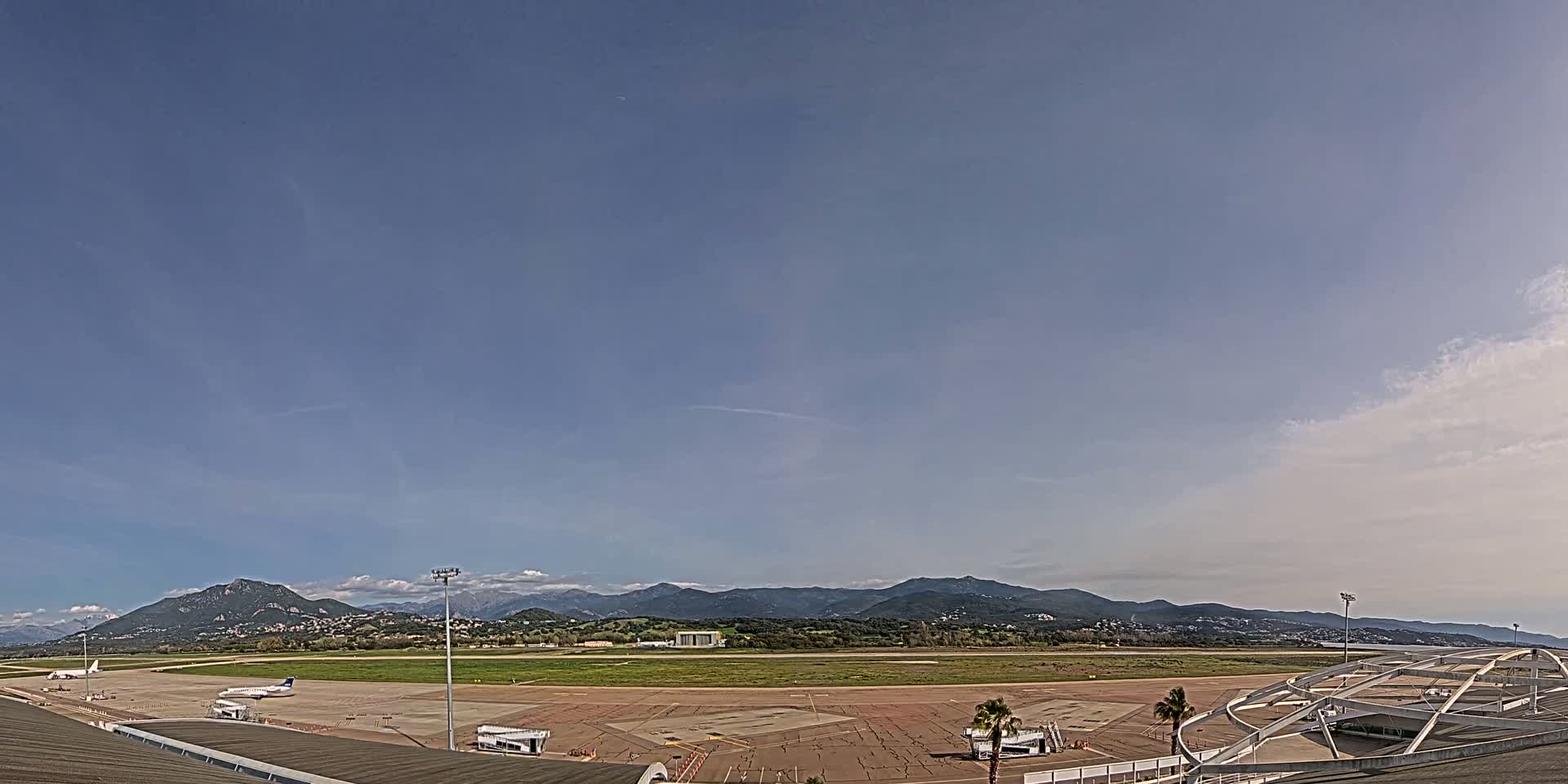 An airport tarmac with several planes, surrounded by grassy fields and mountains under a mostly clear sky.