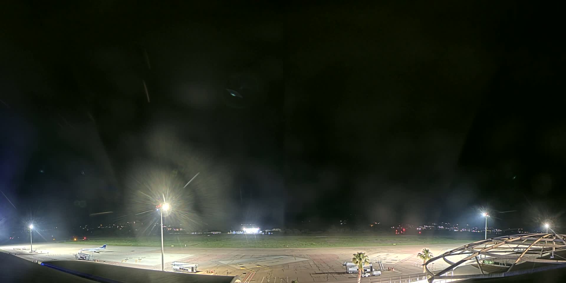 A nighttime view of an airport tarmac with a single airplane, several lights illuminating the area, and a partially visible structure in the foreground.