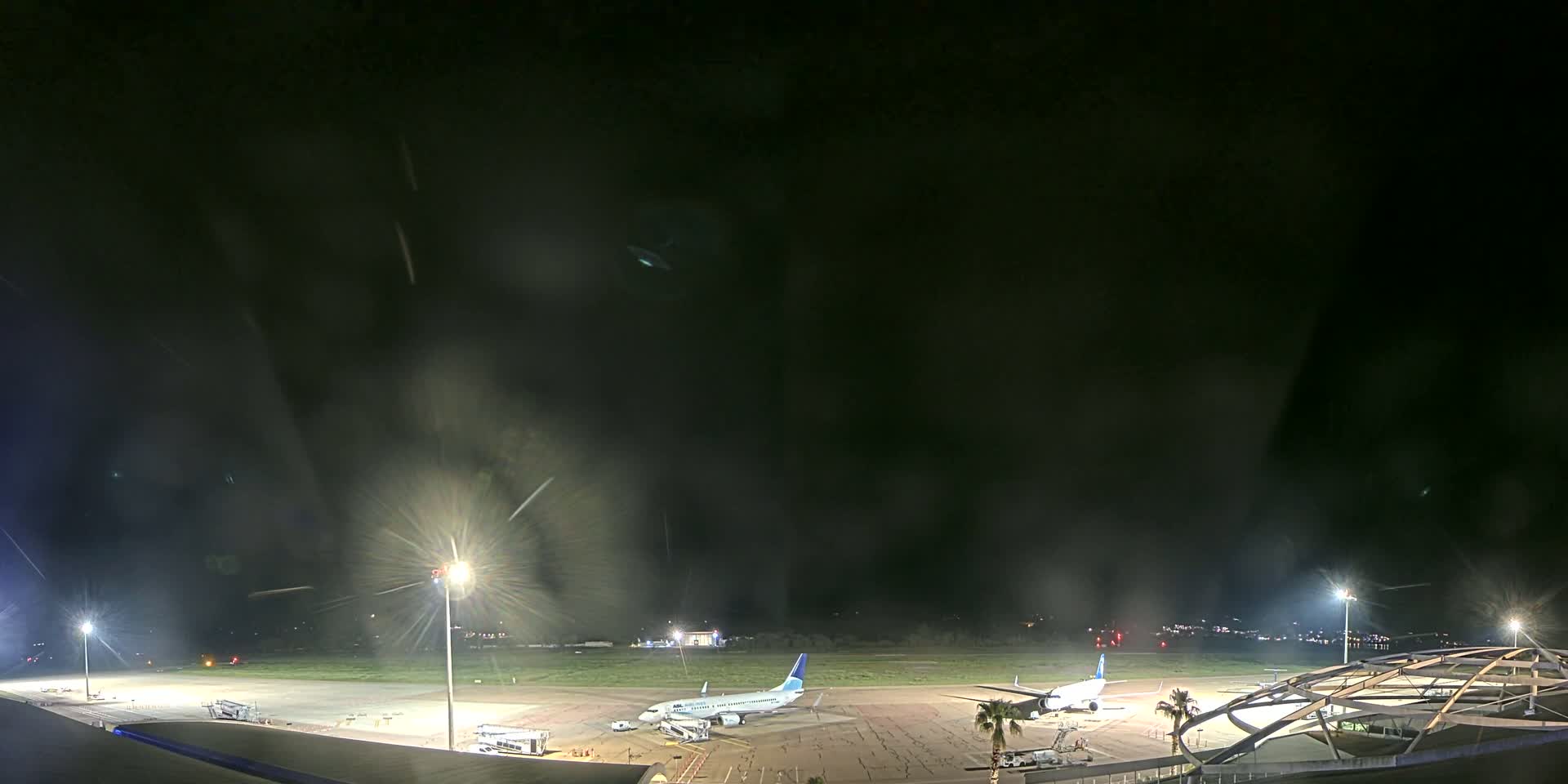 At night, under a dark, possibly slightly hazy sky, two airplanes are parked on a brightly lit airport tarmac.