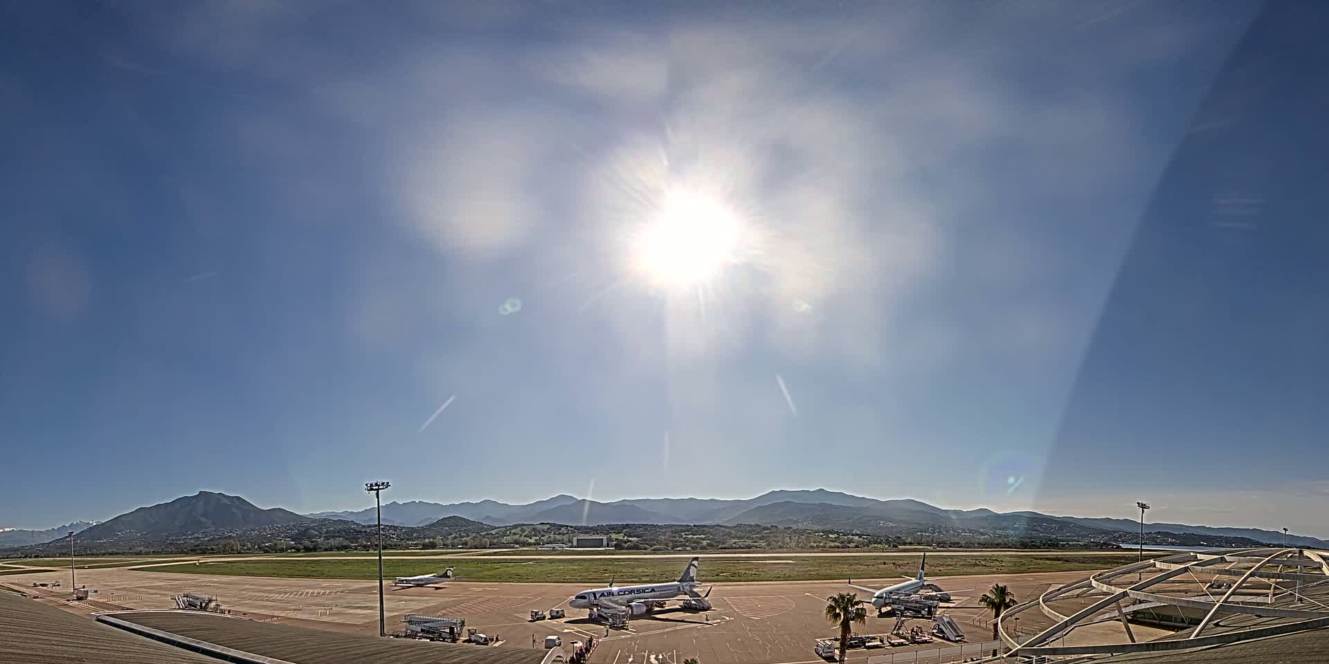 Several airplanes are parked on an airport tarmac under a bright, sunny sky with mountains in the distance.