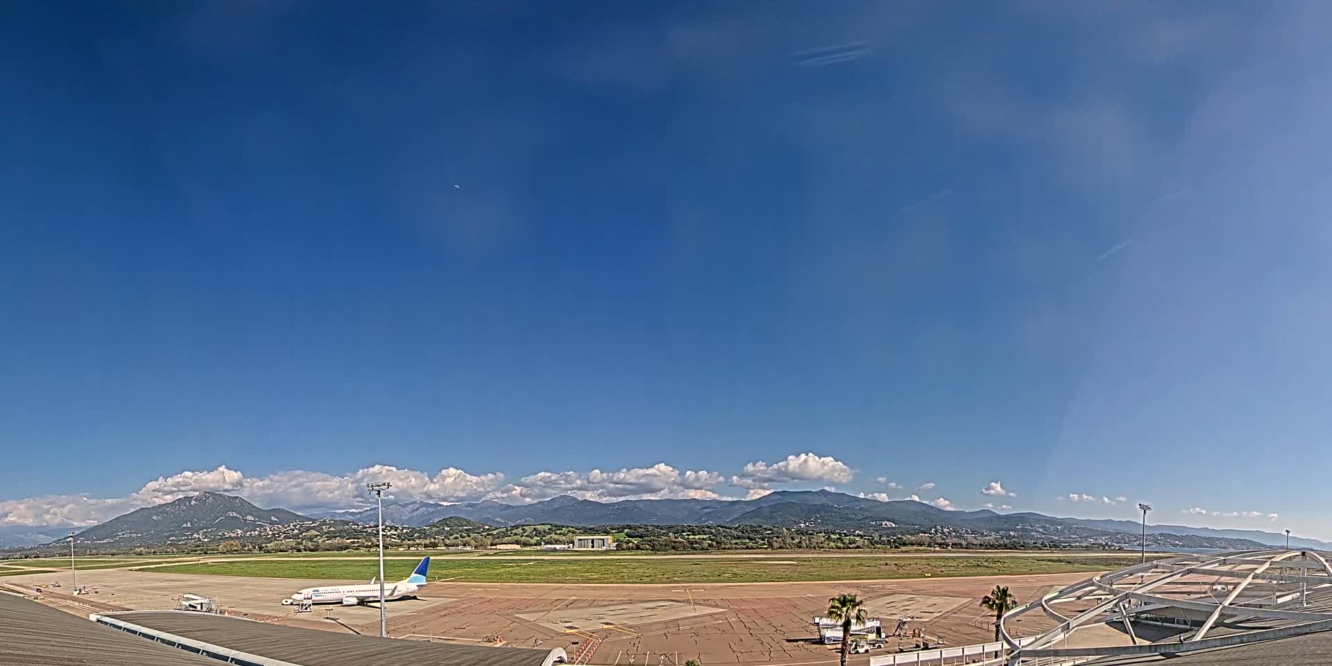 A passenger airplane sits on a tarmac at an airport under a mostly clear blue sky with some cumulus clouds and distant mountains.