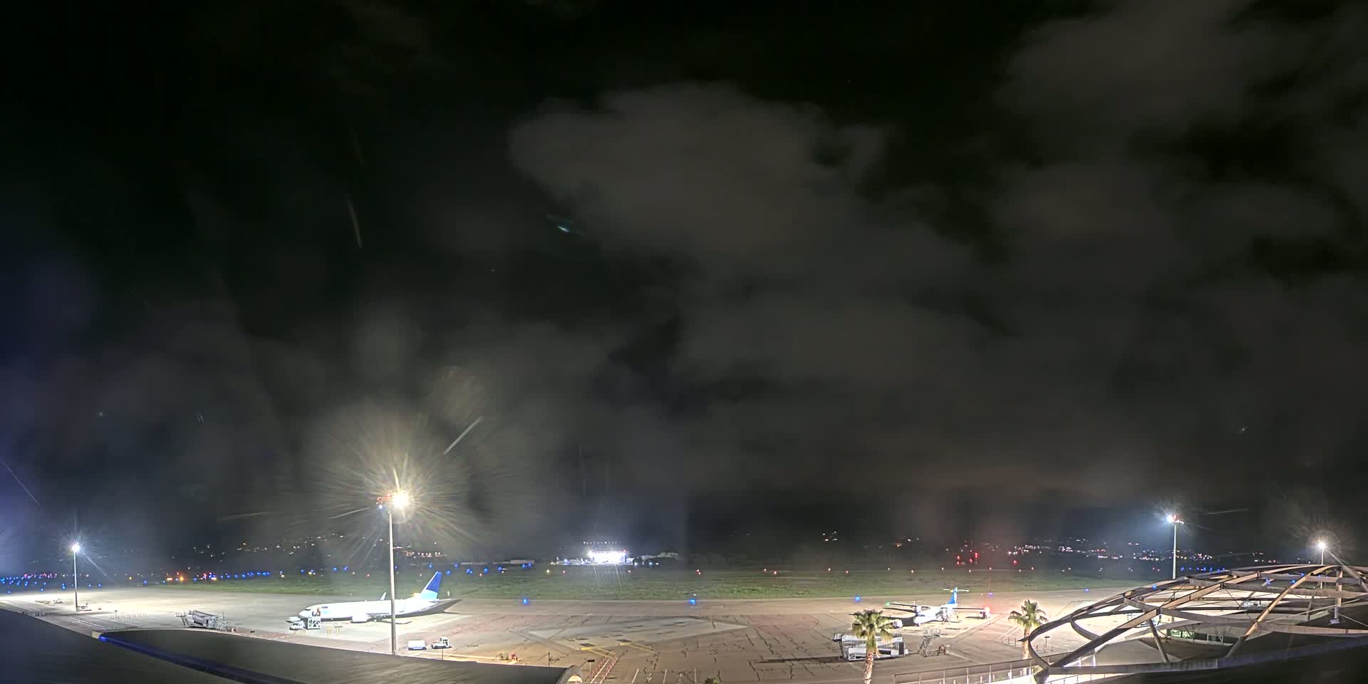 A nighttime view of an airport tarmac with a few airplanes and ground support equipment under a dark, cloudy sky.