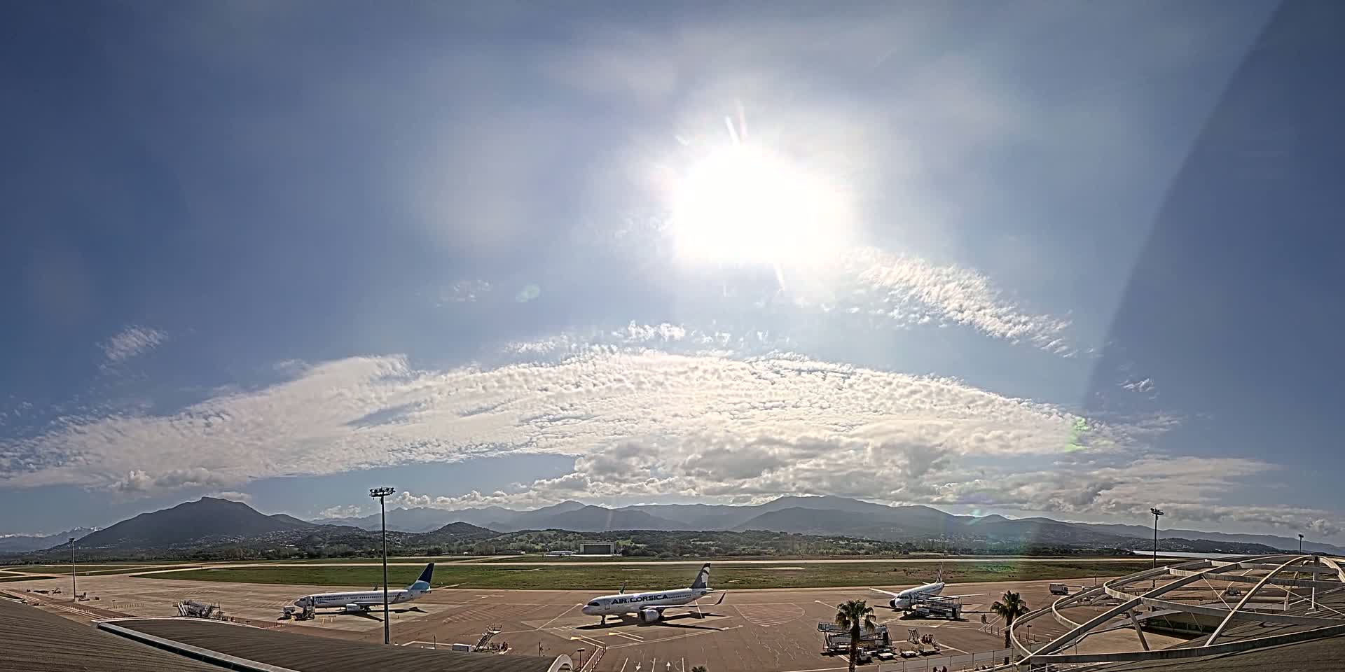 Three airplanes are parked on an airport tarmac under a bright, sunny sky with scattered clouds and distant mountains.