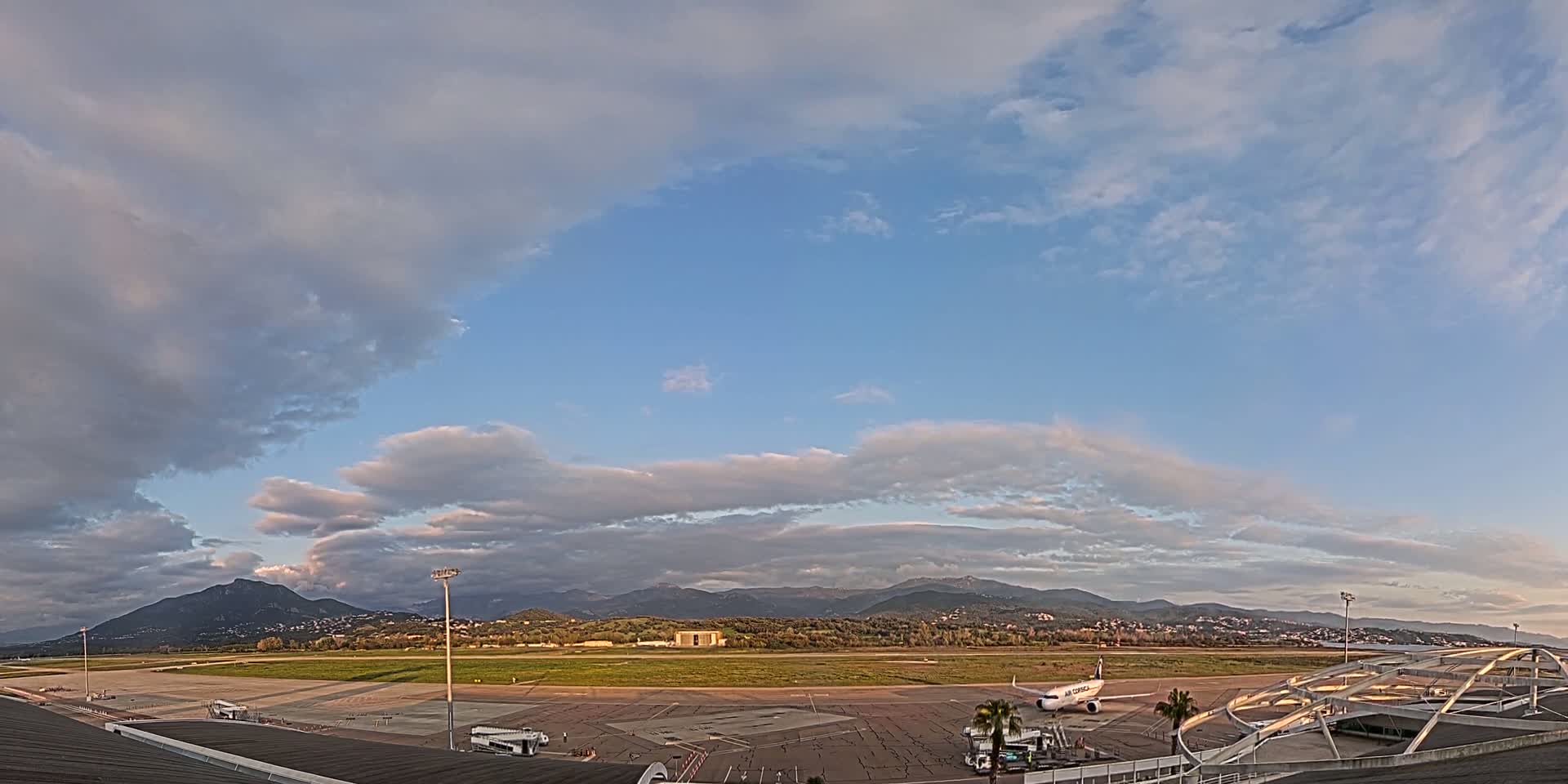 A partly cloudy sky over an airport tarmac with a single airplane parked, mountains in the background.