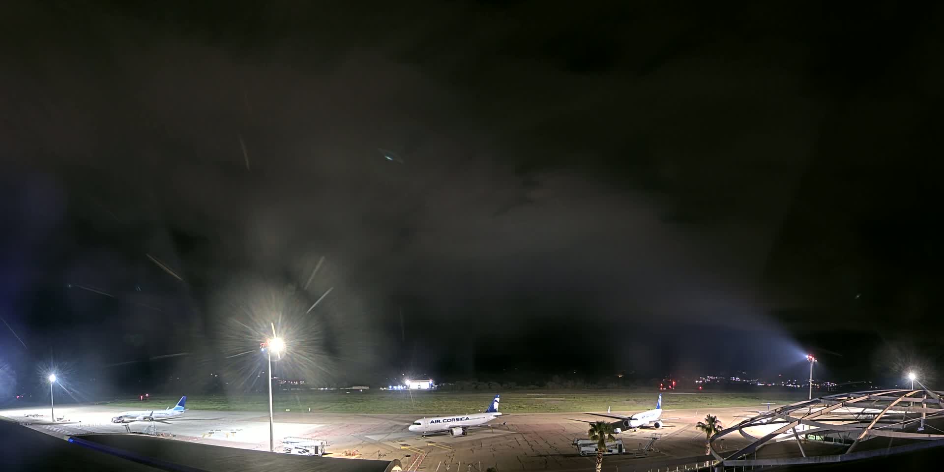 At night, under a dark, cloudy sky, several airplanes are parked on a brightly lit airport tarmac.