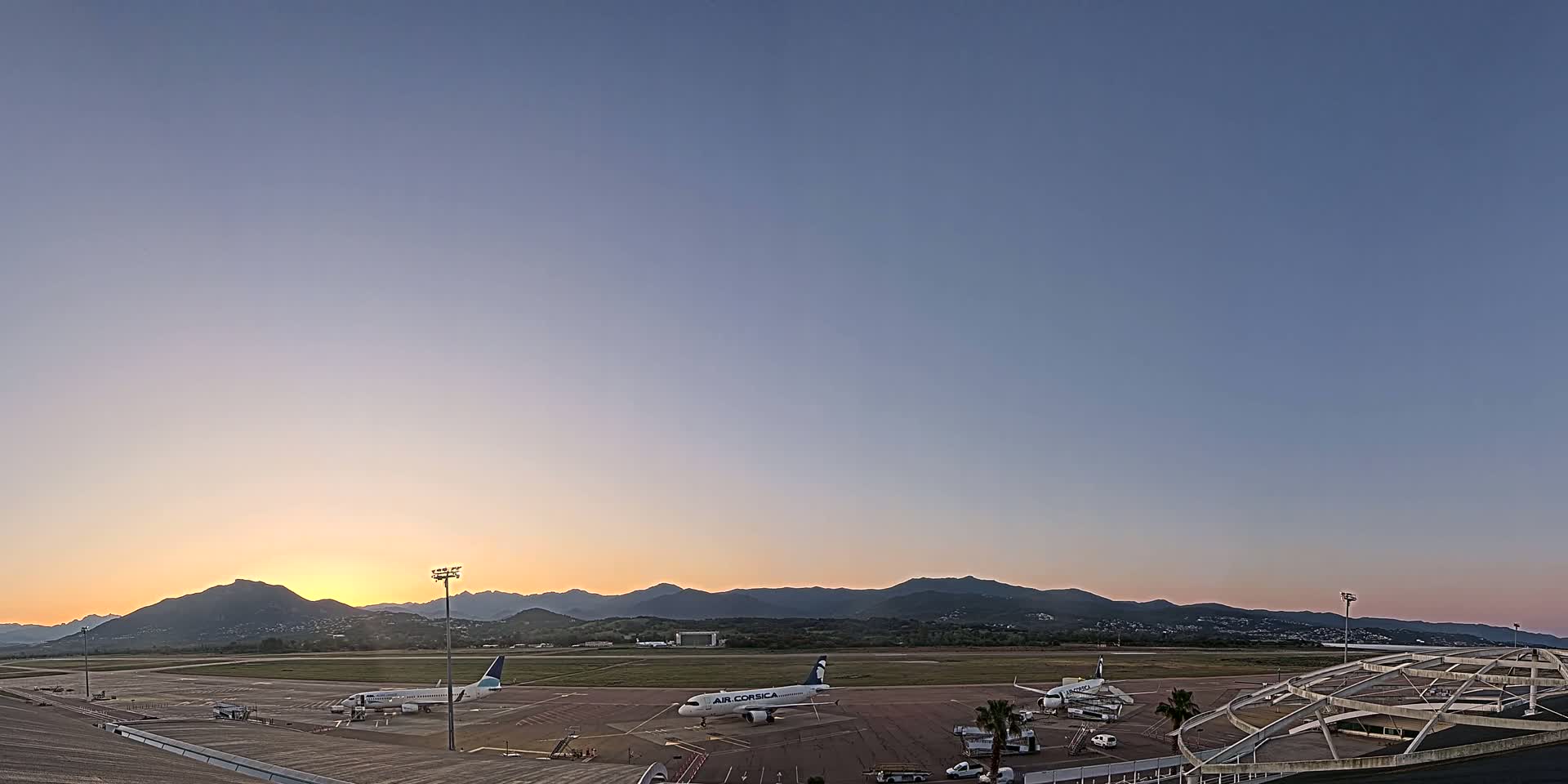 Two airplanes are parked on an airport tarmac at sunrise, with mountains in the background under a clear sky.