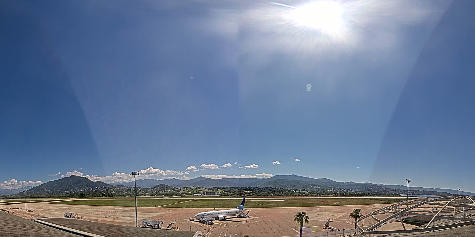 A passenger airplane sits on a tarmac at an airport under a bright, sunny sky with a few scattered clouds and mountains in the distance.