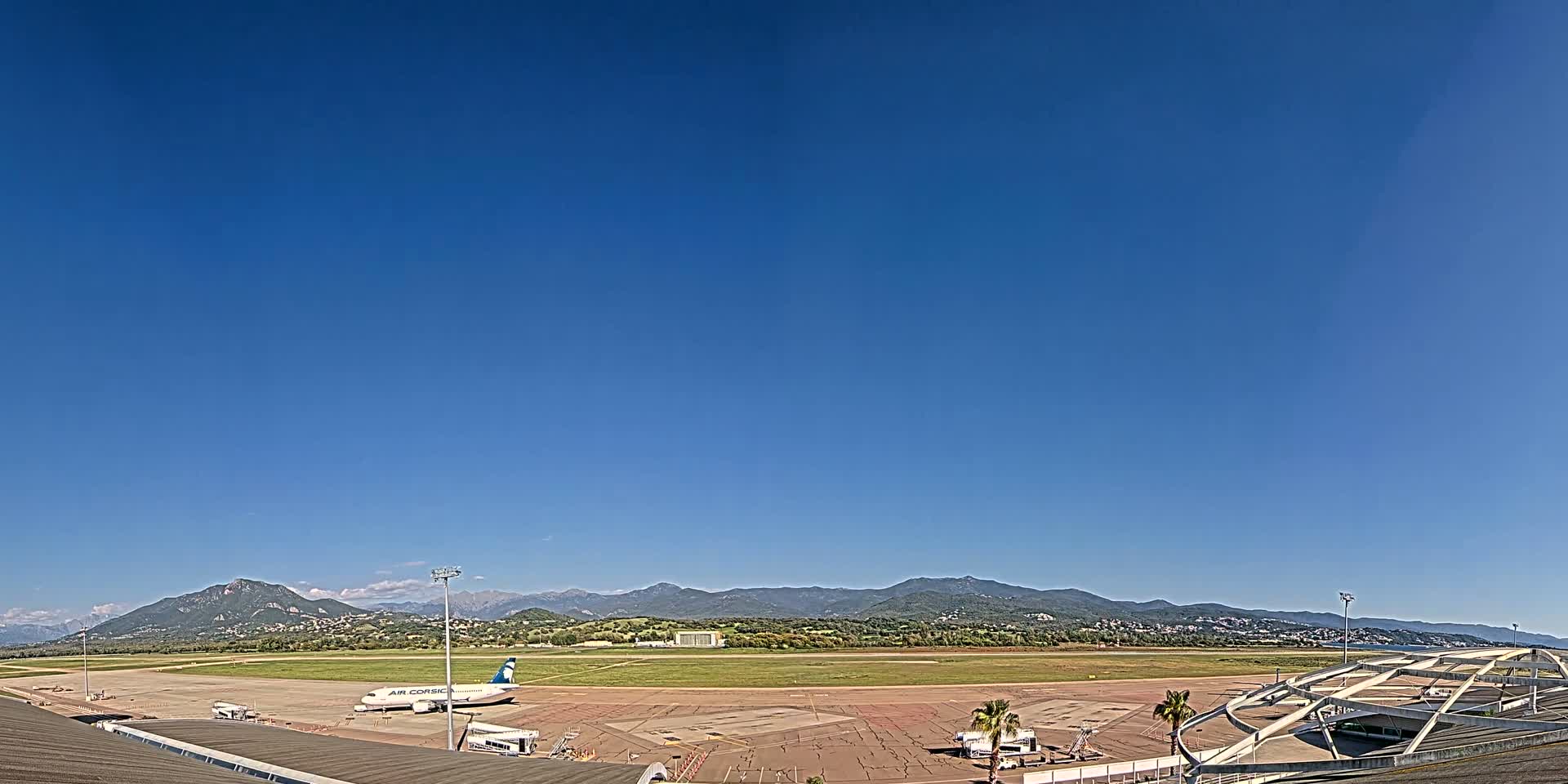 An airplane sits on a tarmac at an airport under a clear blue sky, with mountains visible in the distance.