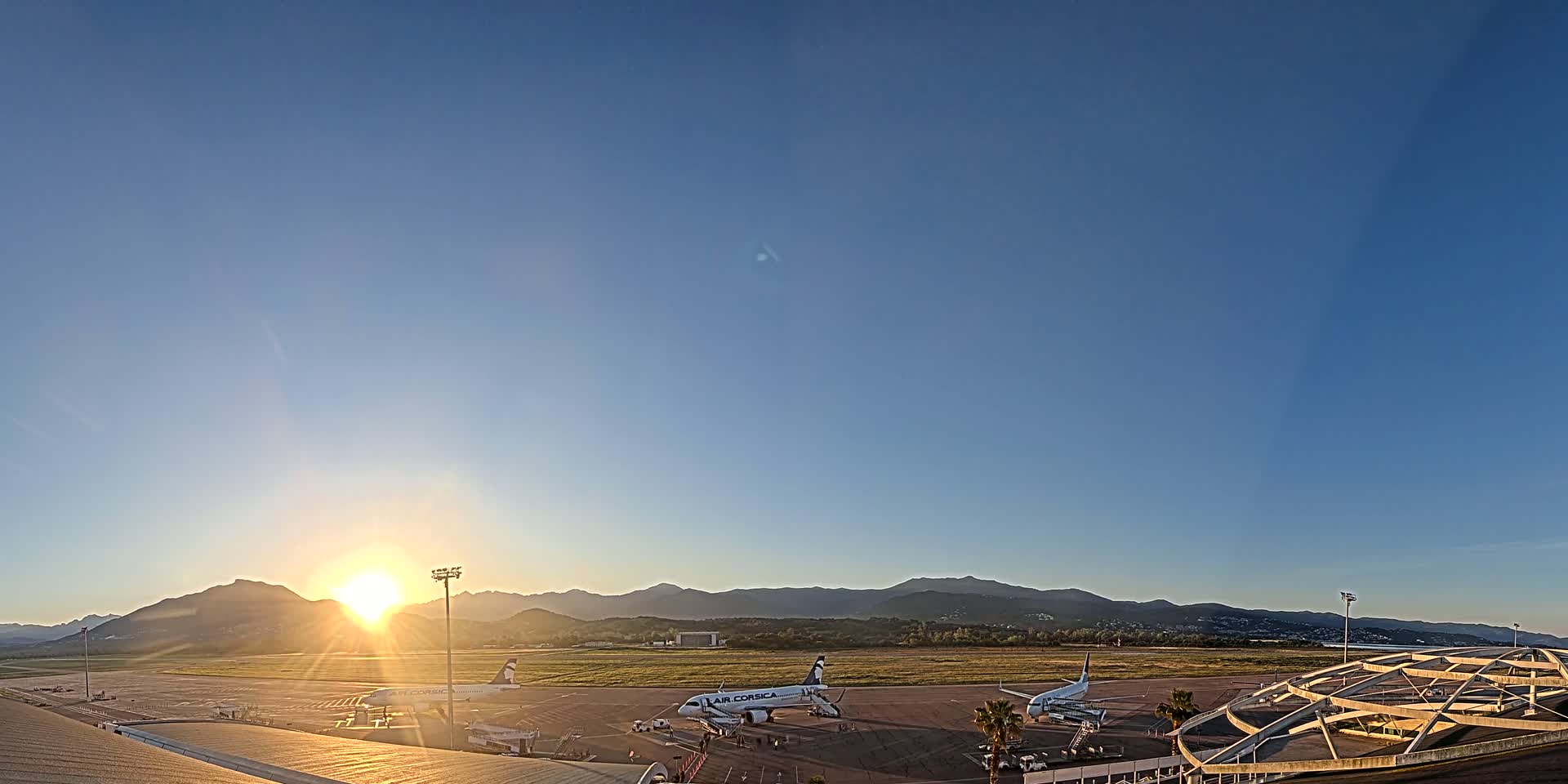 Several airplanes are parked on an airport tarmac at sunrise, with mountains visible in the distance under a clear blue sky.