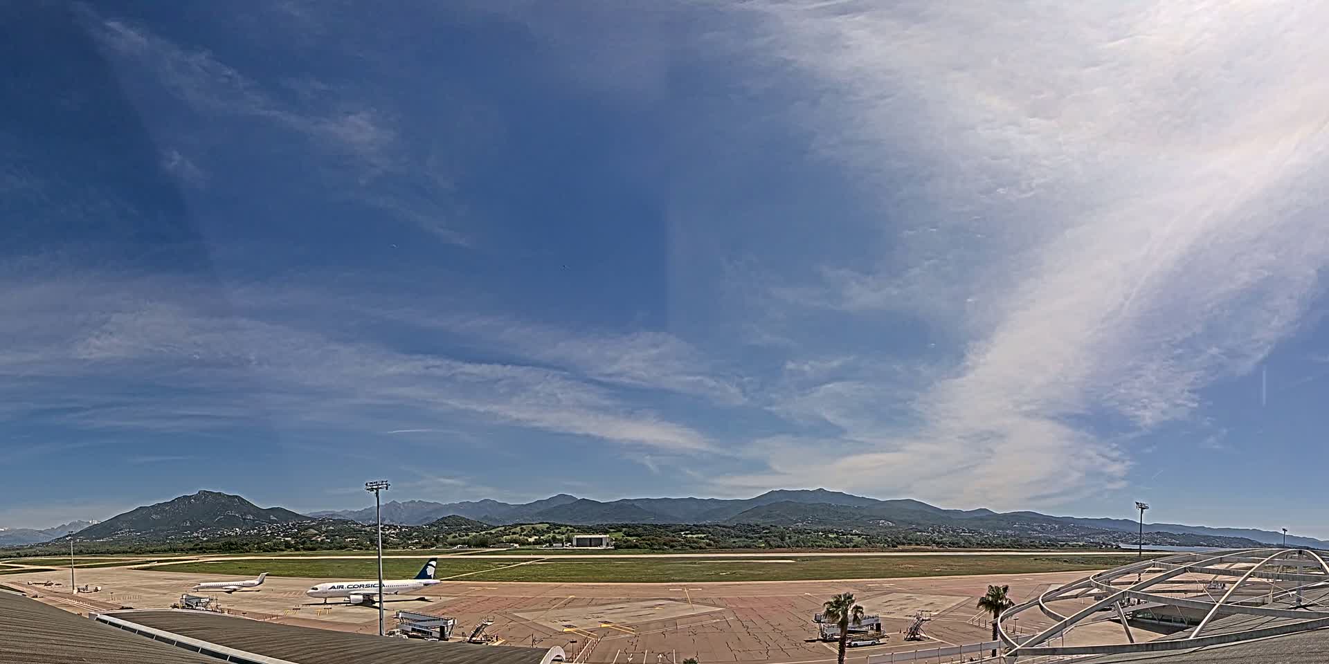 Under a mostly sunny sky with scattered cirrus clouds, an airport tarmac shows several airplanes parked near a terminal building, with mountains visible in the distance.