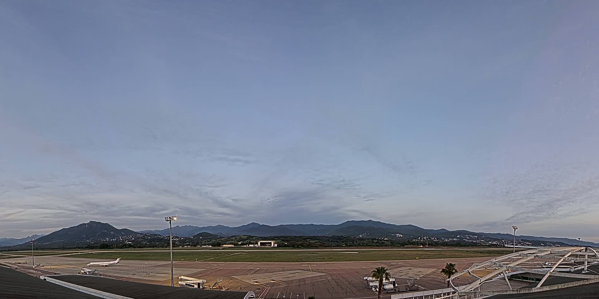 An airport tarmac with a single airplane visible, backed by a range of mountains under a mostly clear, pale sky.