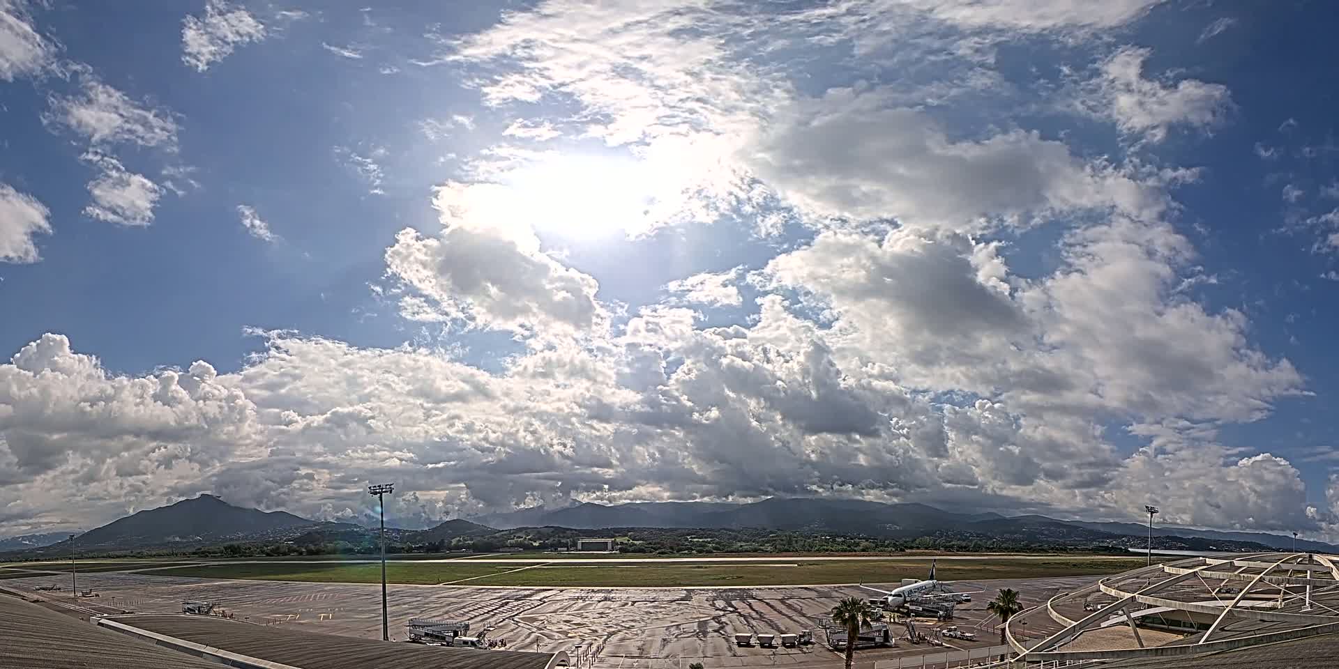 A partly sunny day with fluffy white clouds over an airport tarmac, showing a plane at a gate and mountains in the distance.