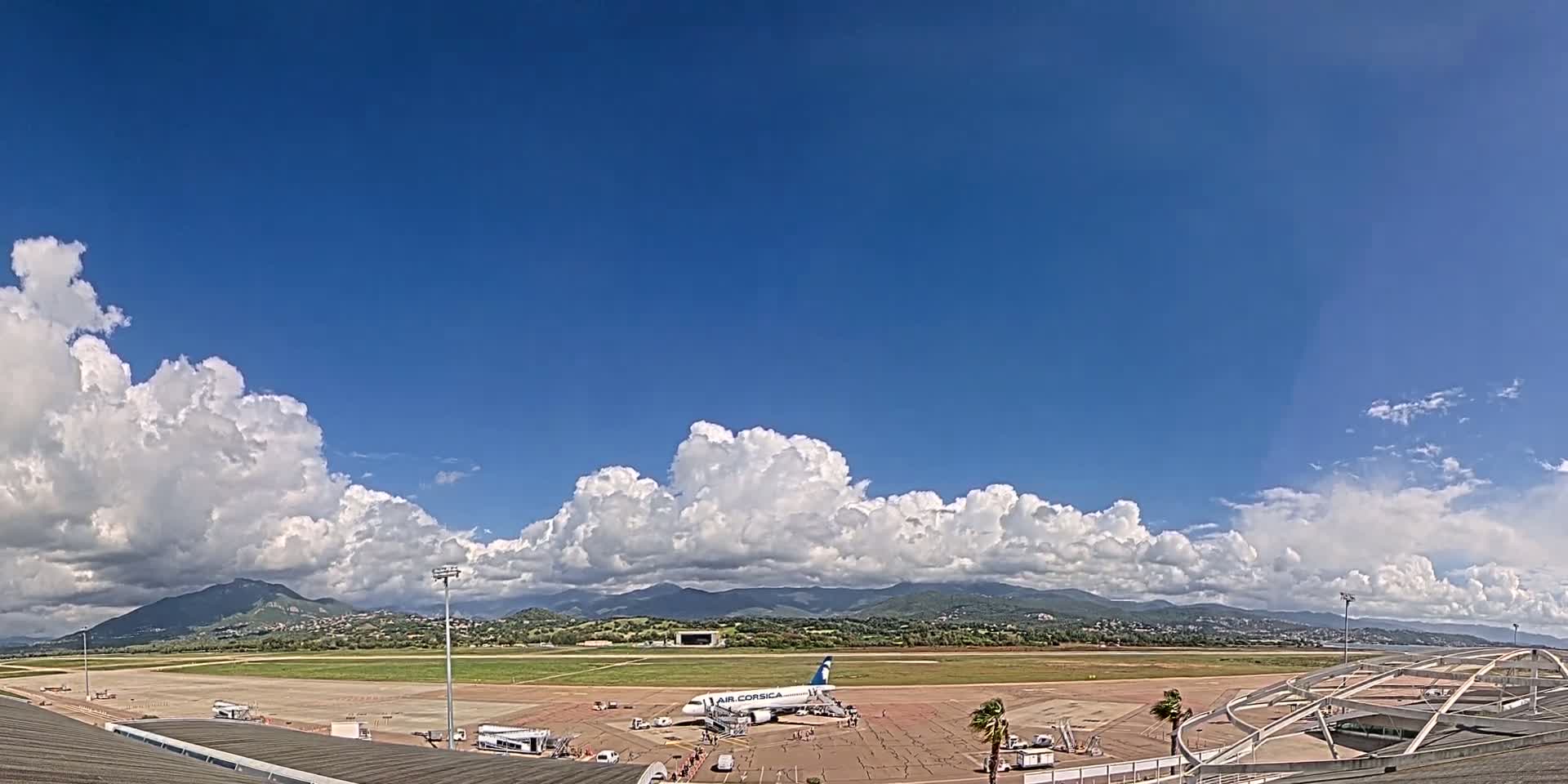A passenger airplane sits on a tarmac at an airport under a mostly sunny sky with puffy cumulus clouds and mountains in the distance.