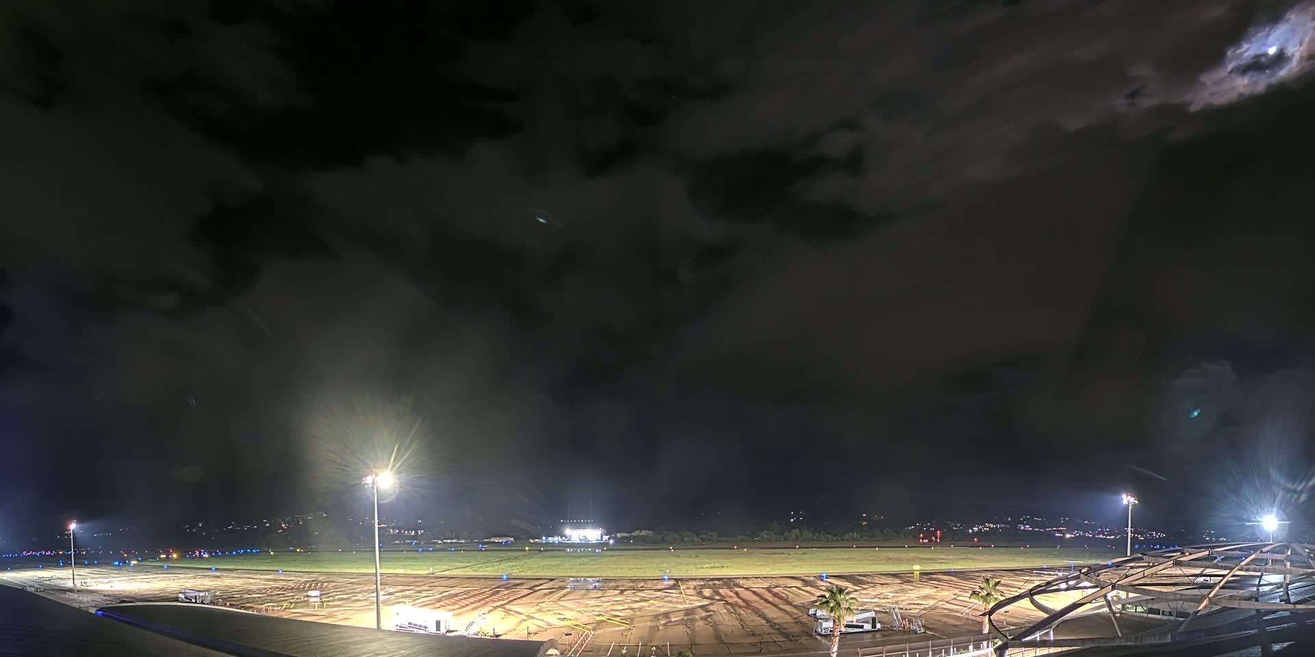 A nighttime view of an airport tarmac under a partly cloudy sky, illuminated by bright lights.