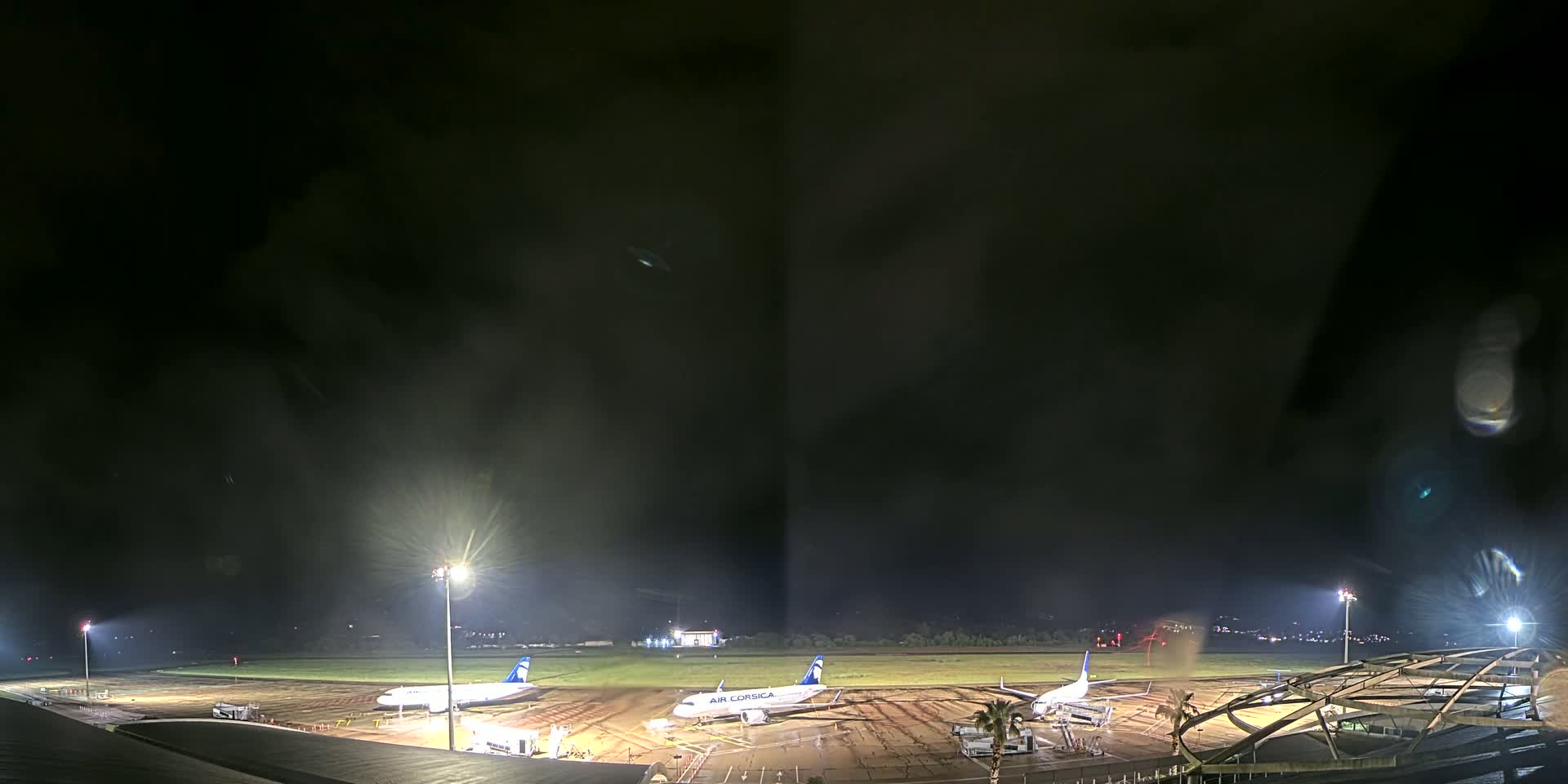 Three airplanes are parked on a dark, wet airport tarmac at night under bright floodlights.
