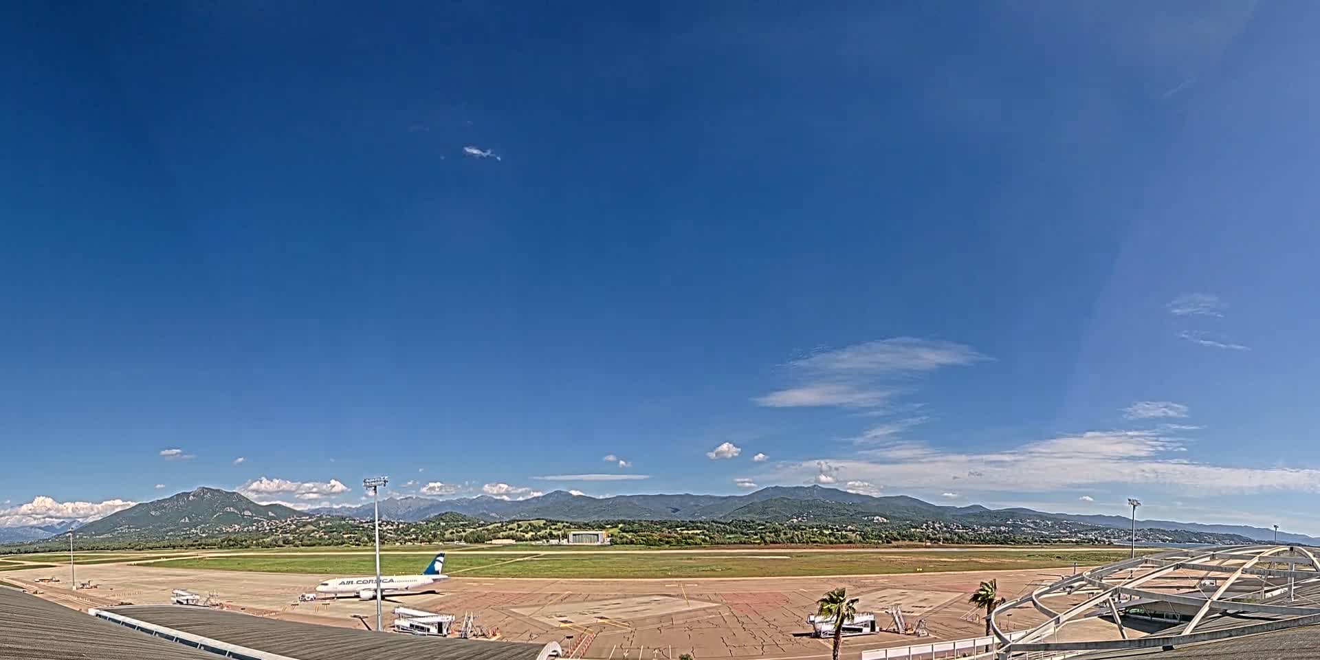 A mostly sunny day at an airport with a single airplane parked on the tarmac, mountains in the distance, and some clouds in the sky.