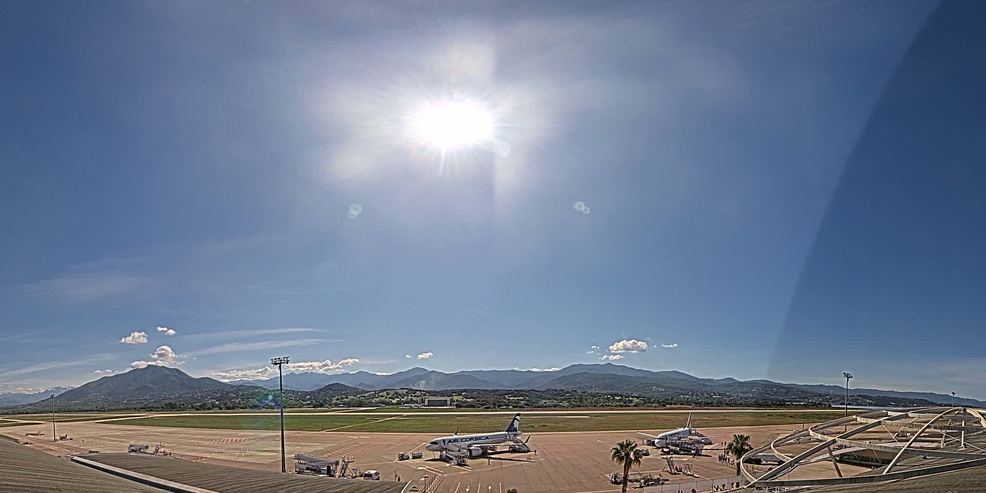 A sunny day at an airport shows two airplanes on the tarmac with mountains visible in the distance.