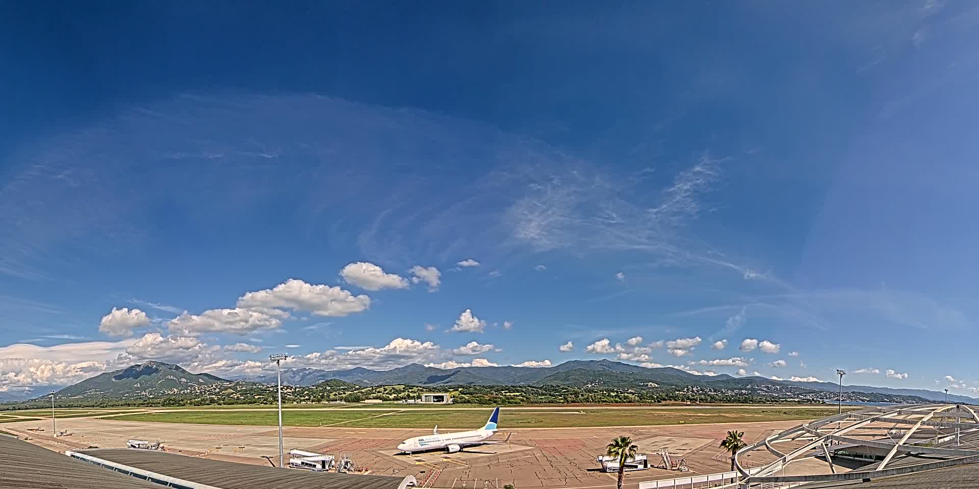 A passenger airplane sits on a tarmac at an airport under a mostly sunny sky with scattered cumulus clouds and mountains in the distance.