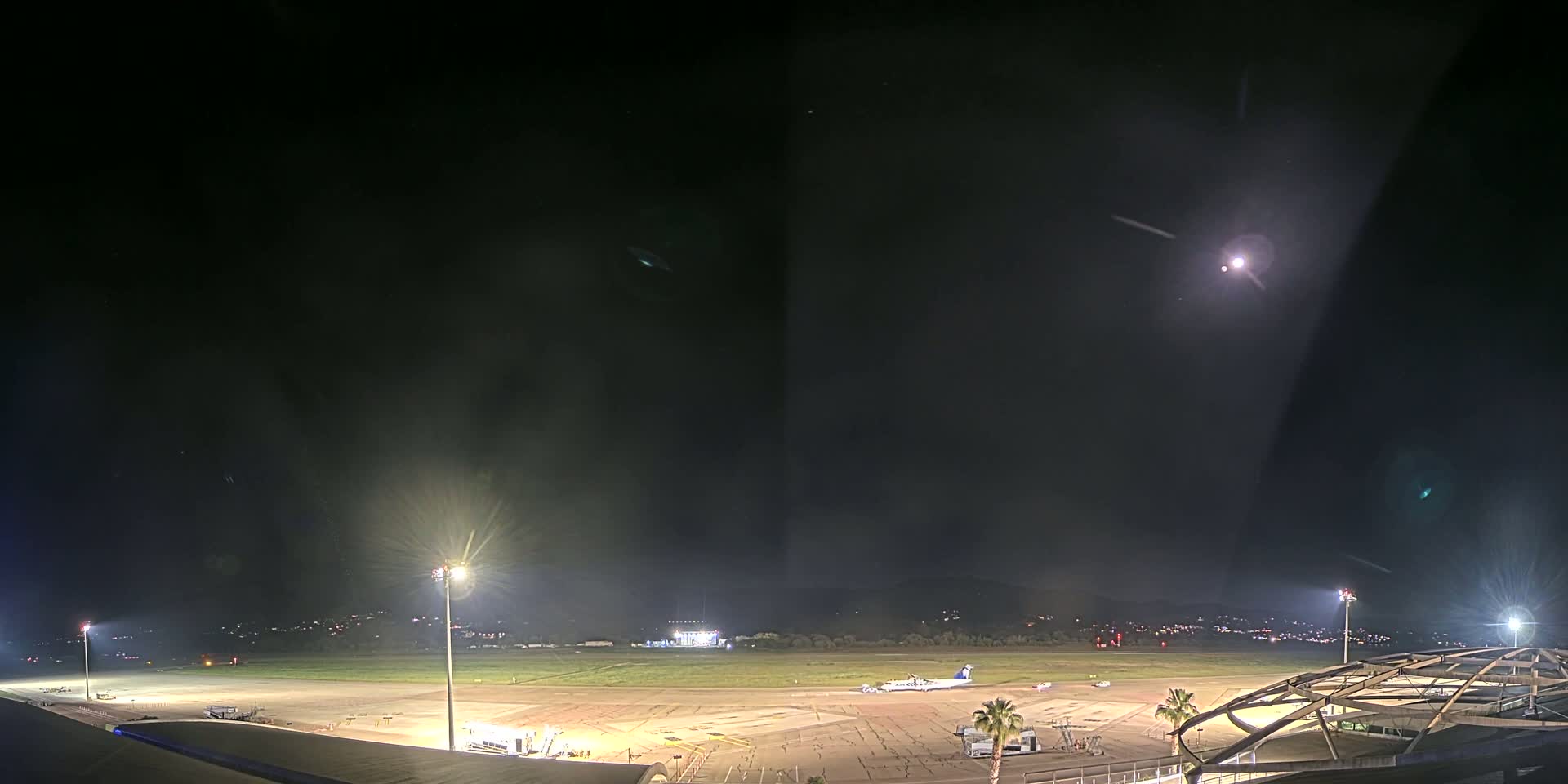A brightly lit airport tarmac at night, under a clear sky with a visible moon, shows a single airplane parked near a terminal building.