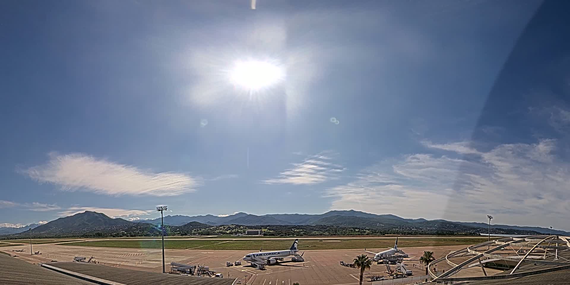 A sunny day at an airport shows two airplanes on the tarmac with mountains in the background and a few wispy clouds in the sky.