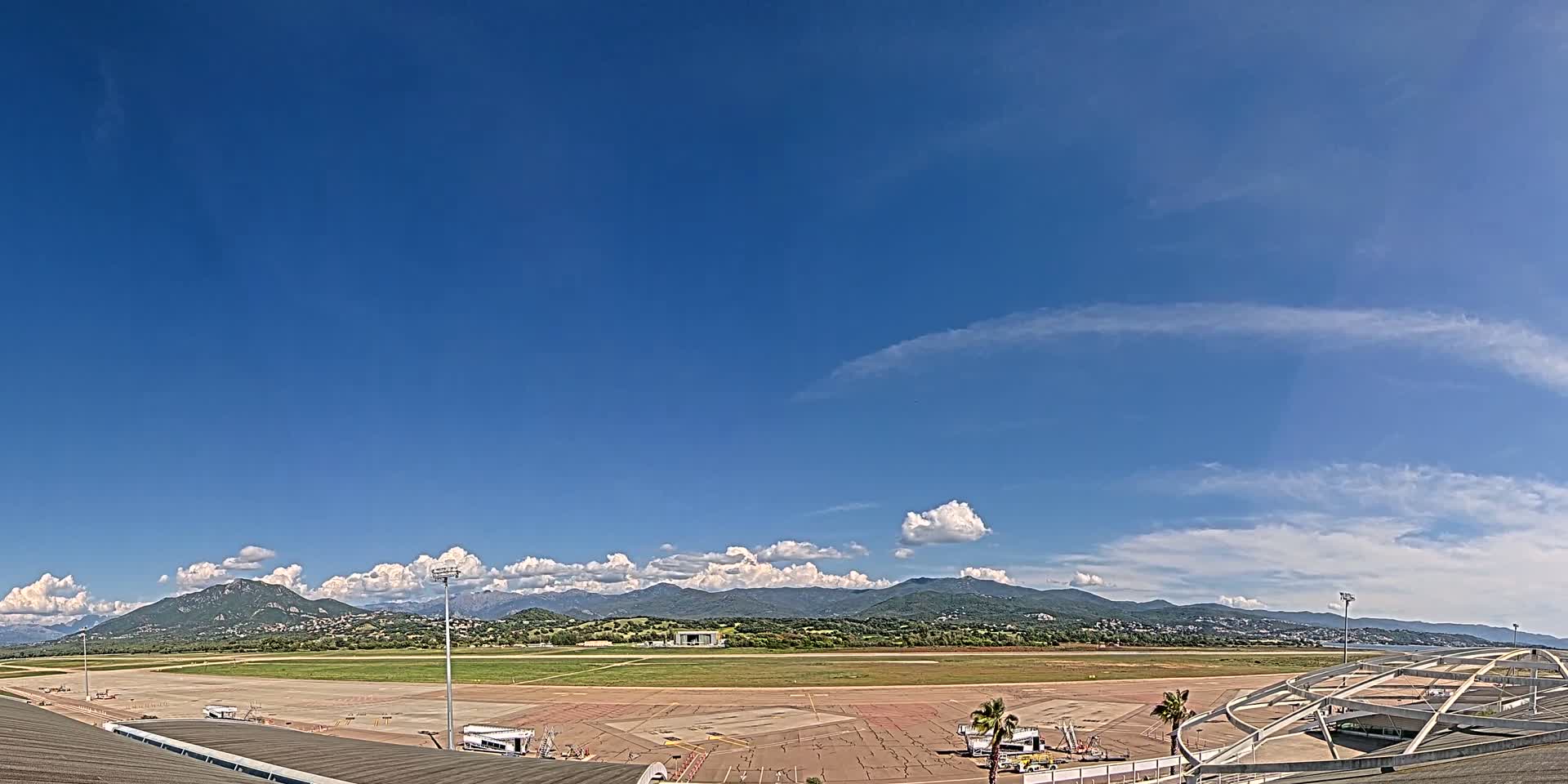 A mostly sunny day shows an airport tarmac with a runway and distant mountains under a blue sky with scattered cumulus clouds.