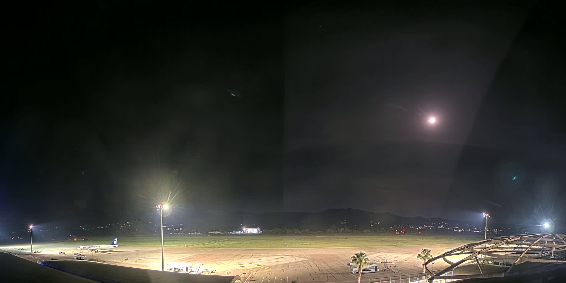 A brightly lit airport tarmac at night under a mostly clear sky with a full moon, showing a single airplane parked near a terminal building and surrounding landscape.