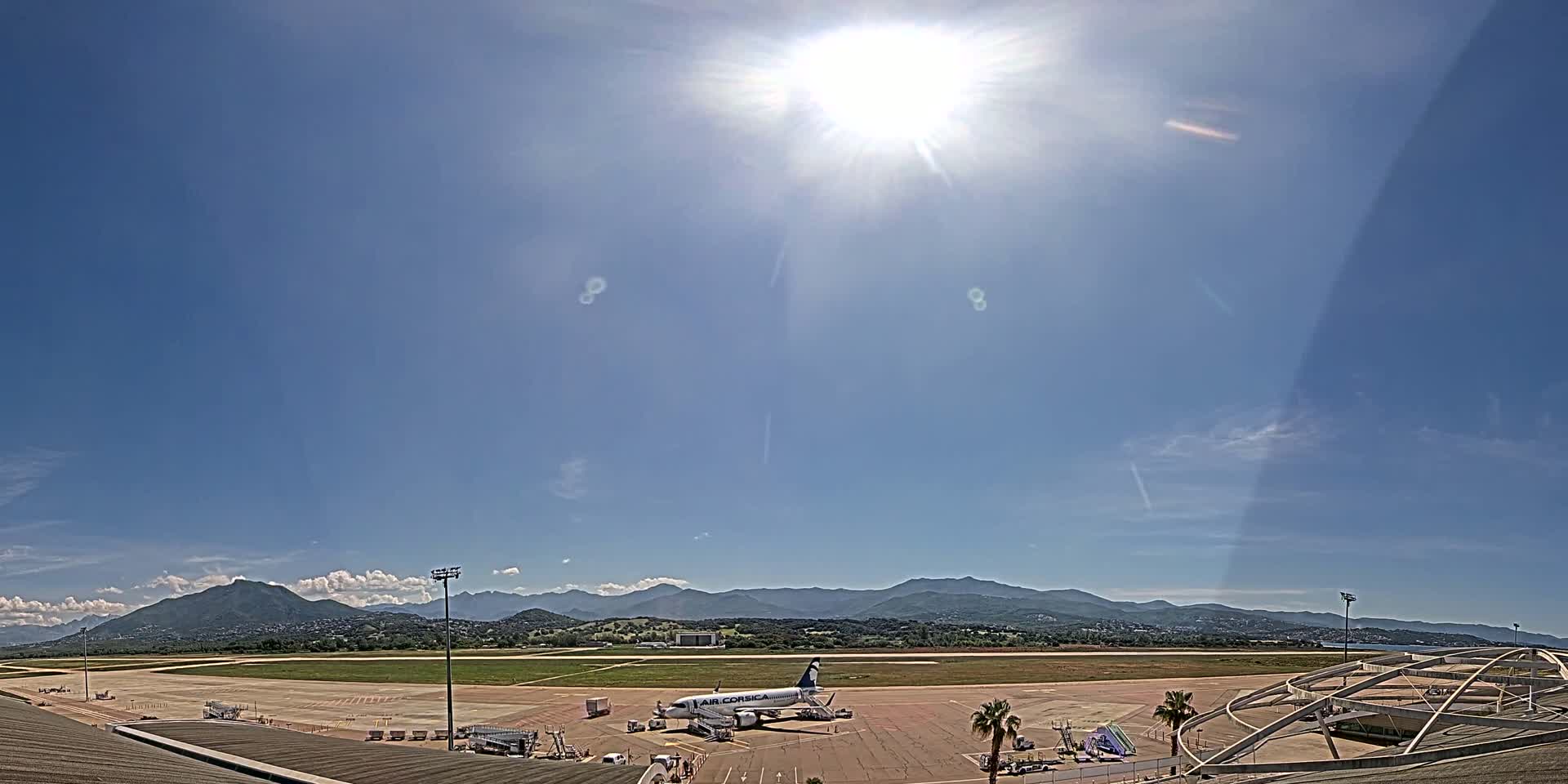 A sunny day at an airport shows a single airplane parked on the tarmac with mountains in the background.