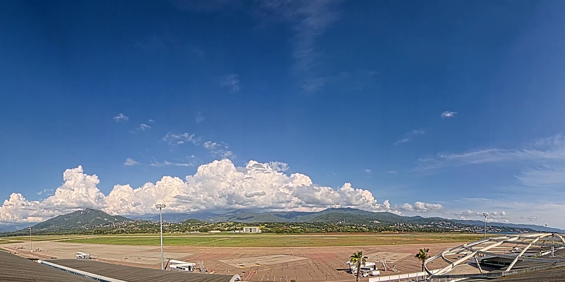 A mostly sunny day with puffy white cumulus clouds over a large airport tarmac and runway, backed by distant mountains.