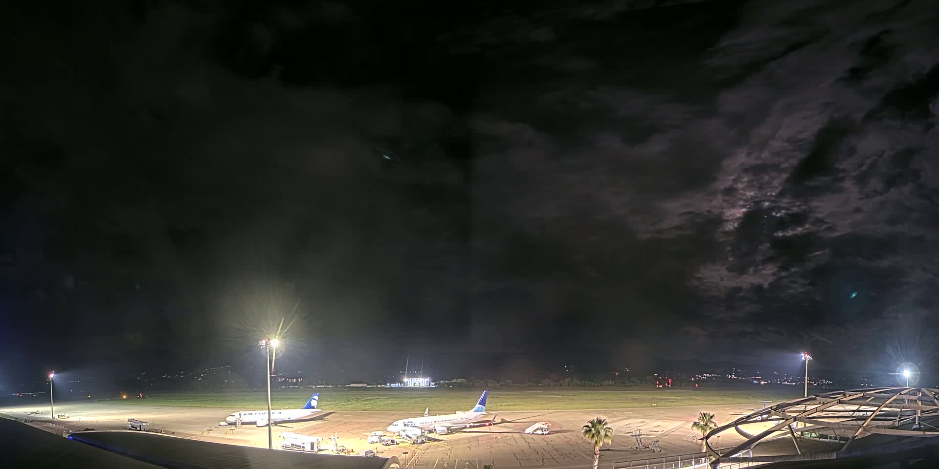 Under a dark, cloudy night sky, two airplanes are parked on a brightly lit airport tarmac.
