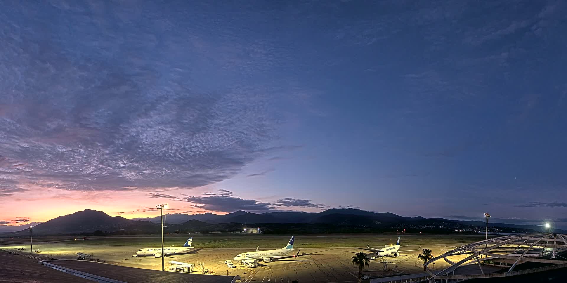Three airplanes are parked on an airport tarmac at twilight under a mostly clear sky with thin clouds, mountains visible in the distance.