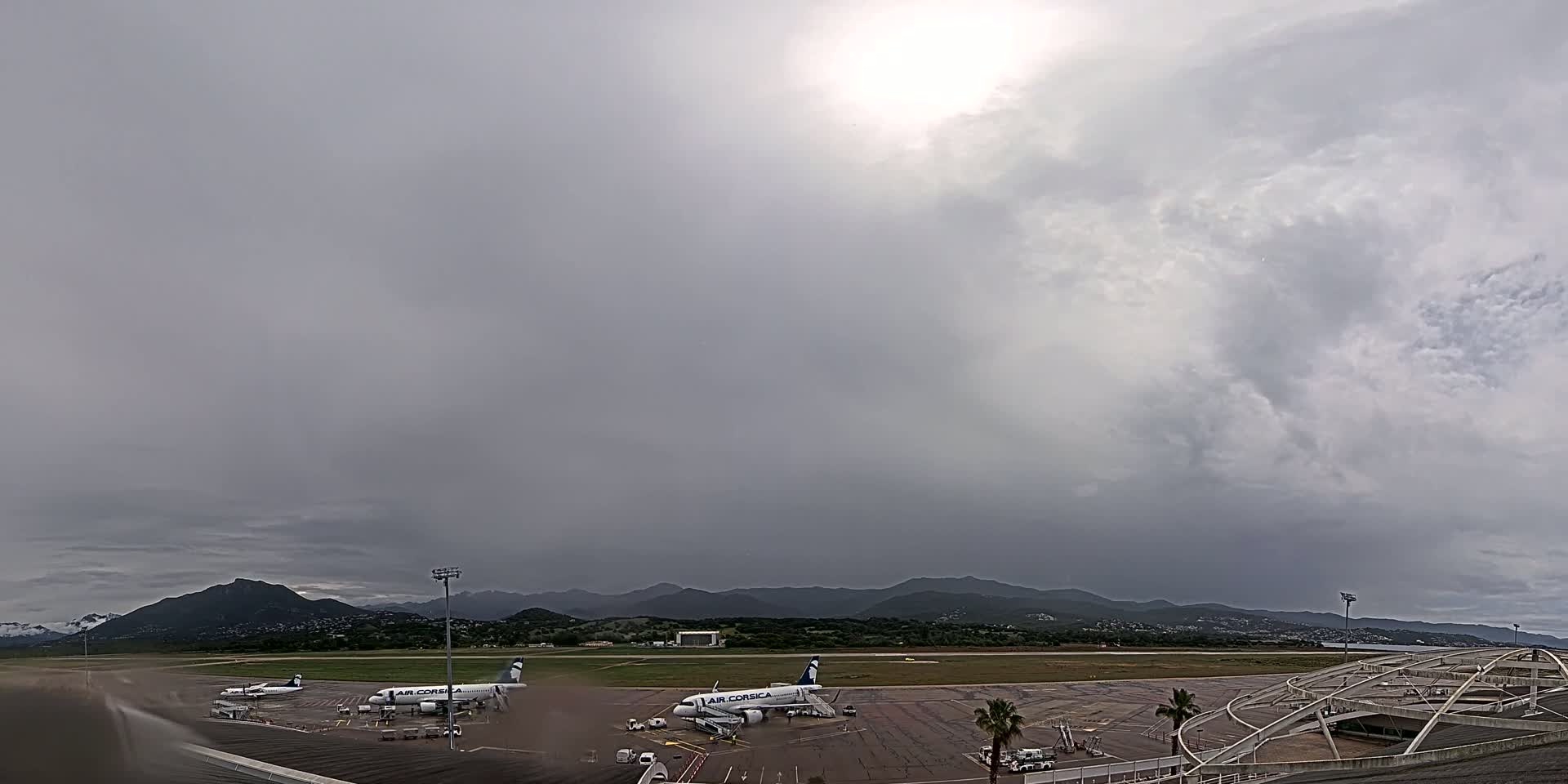Two airplanes are parked on an airport tarmac under an overcast sky, with mountains visible in the distance.