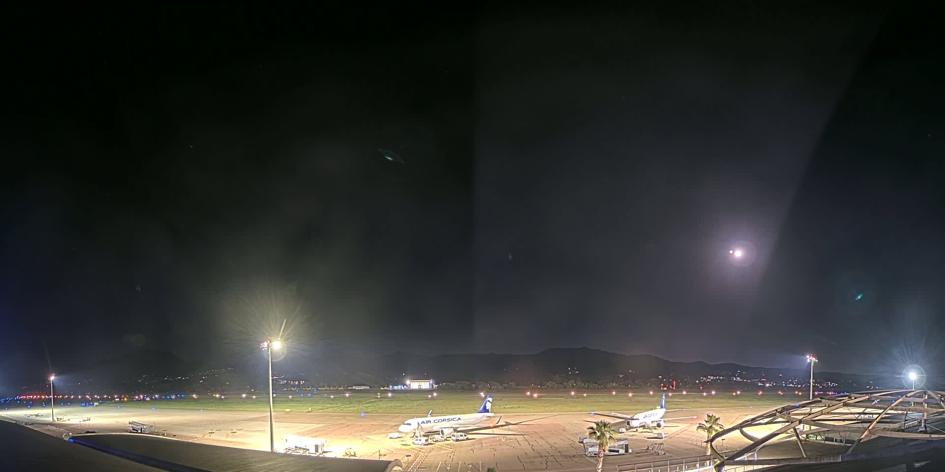 At night, under a mostly clear sky illuminated by a bright moon, two airplanes sit on a tarmac at an airport surrounded by lights, with hills visible in the distance.