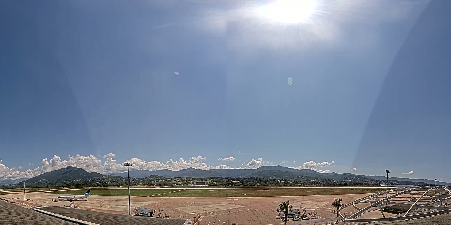 A sunny day at an airport with a large airplane parked on the tarmac, mountains in the background, and some clouds in the sky.