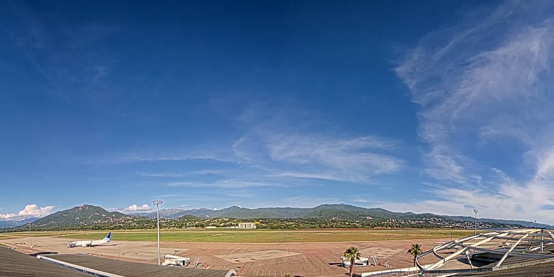 A mostly clear blue sky with wispy clouds overlooks an airport tarmac with a single airplane parked, adjacent to mountains in the distance.