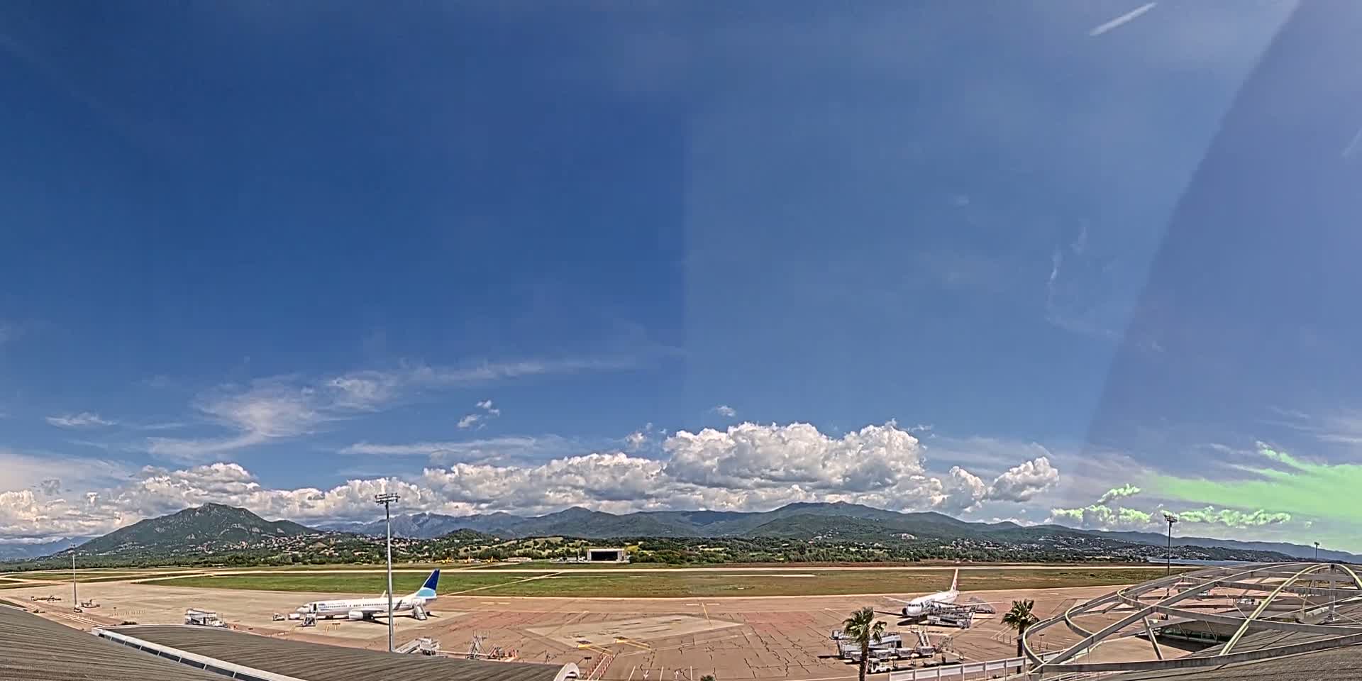A partly cloudy sky overlooks an airport tarmac with several airplanes parked near a terminal building and mountains in the distance.