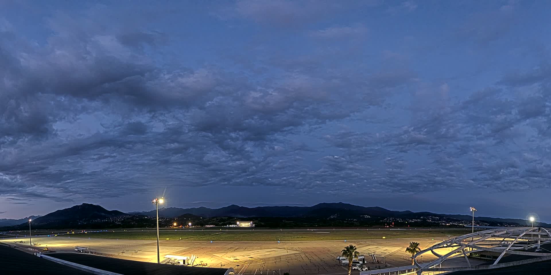 An illuminated airport tarmac at twilight under a mostly cloudy sky, with mountains in the distance.