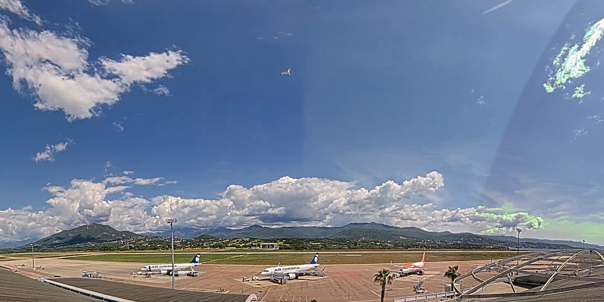 Three airplanes are parked on an airport tarmac under a partly cloudy blue sky, with mountains visible in the distance.