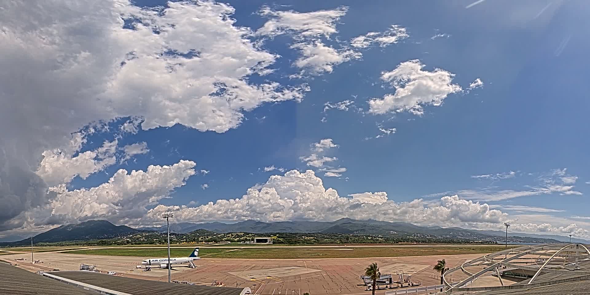 A partly cloudy sky over an airport tarmac with a single airplane and distant mountains.