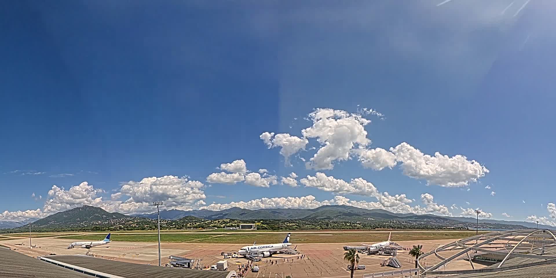 Several airplanes are parked on a tarmac at an airport under a mostly sunny sky with scattered cumulus clouds, mountains visible in the distance.