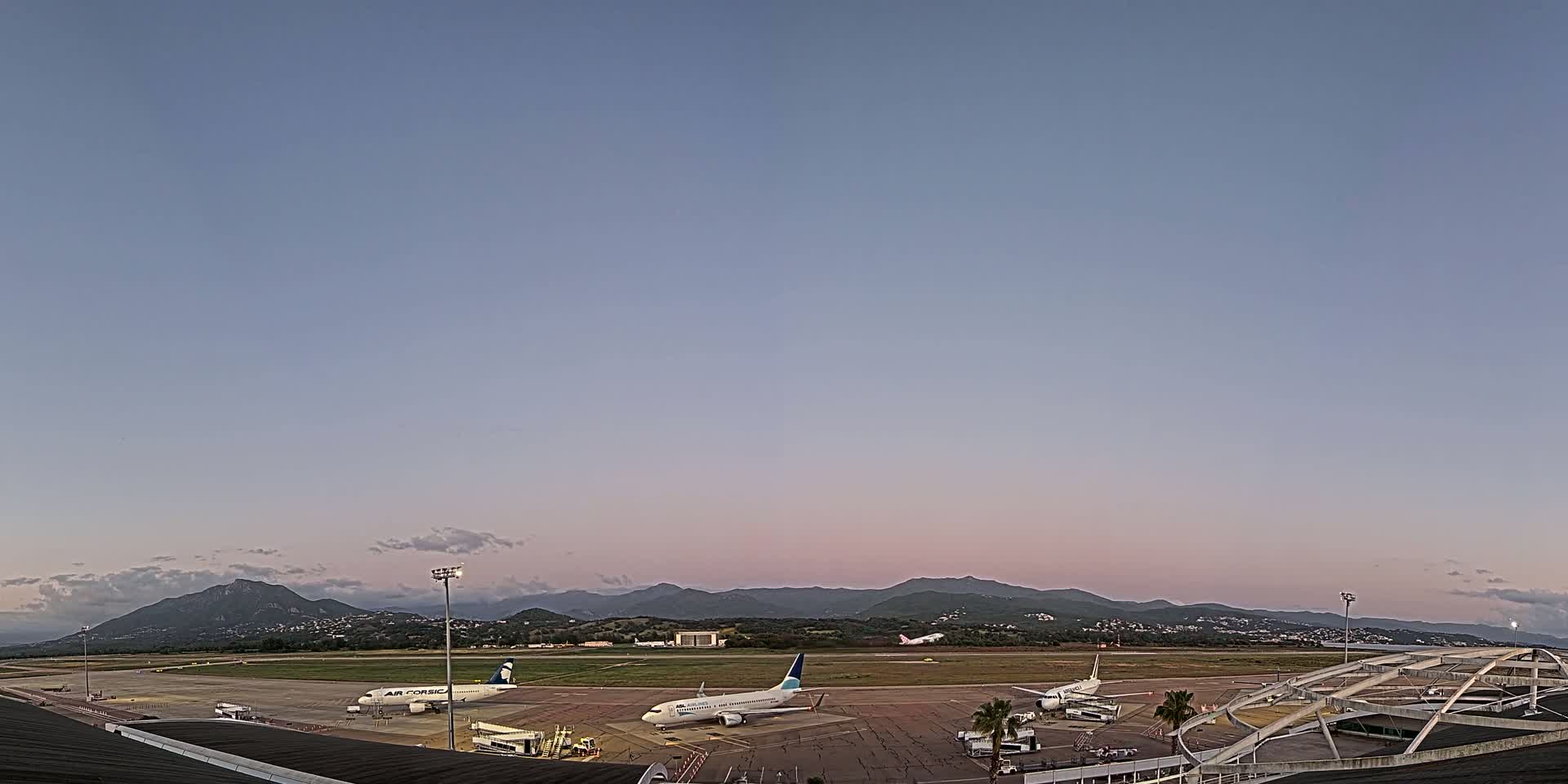 Several airplanes are parked on an airport tarmac under a twilight sky, with mountains visible in the distance.