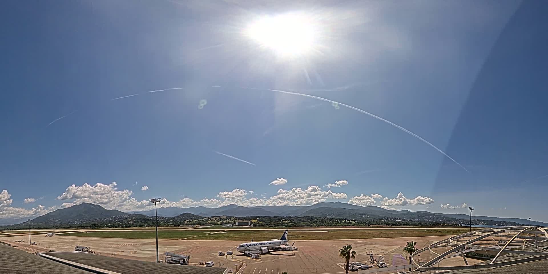 A passenger airplane sits on an airport tarmac under a bright, sunny sky with scattered clouds and distant mountains.