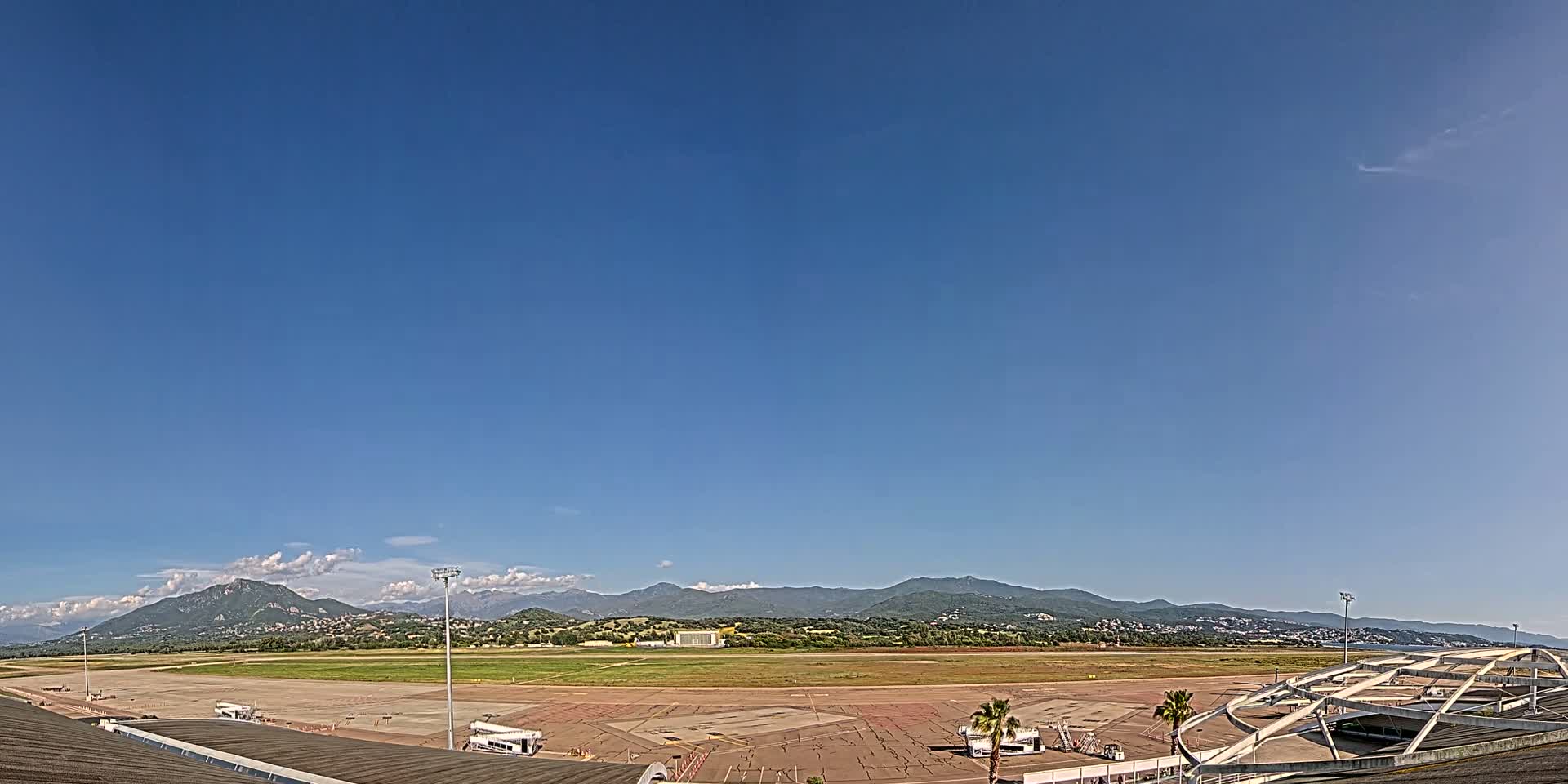 A mostly sunny day reveals an airport tarmac, apron, and runway, with mountains visible in the distance.