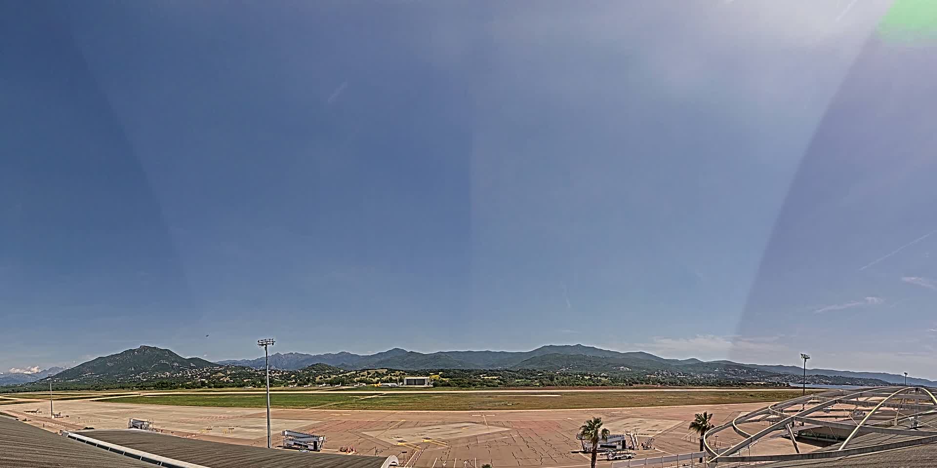 A mostly sunny day shows an airport tarmac with a runway and several aircraft parked, backed by a mountain range under a clear blue sky.