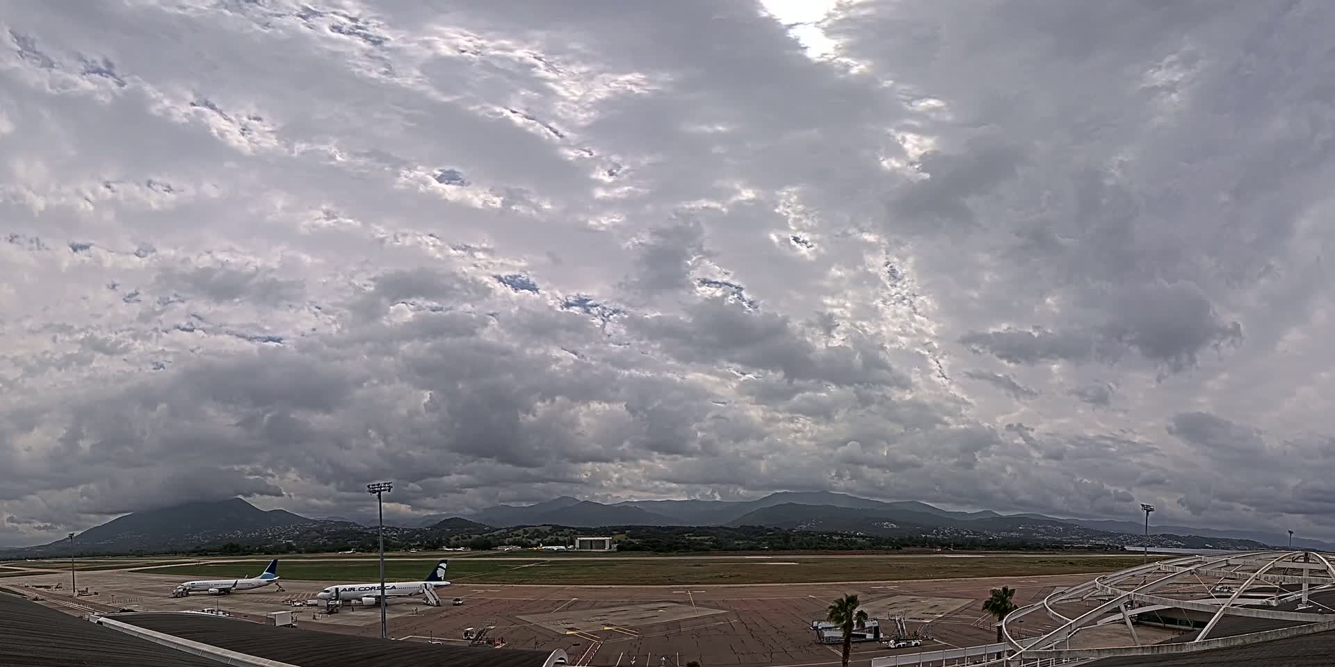 Under a mostly cloudy sky, two airplanes are parked on an airport tarmac with mountains visible in the distance.