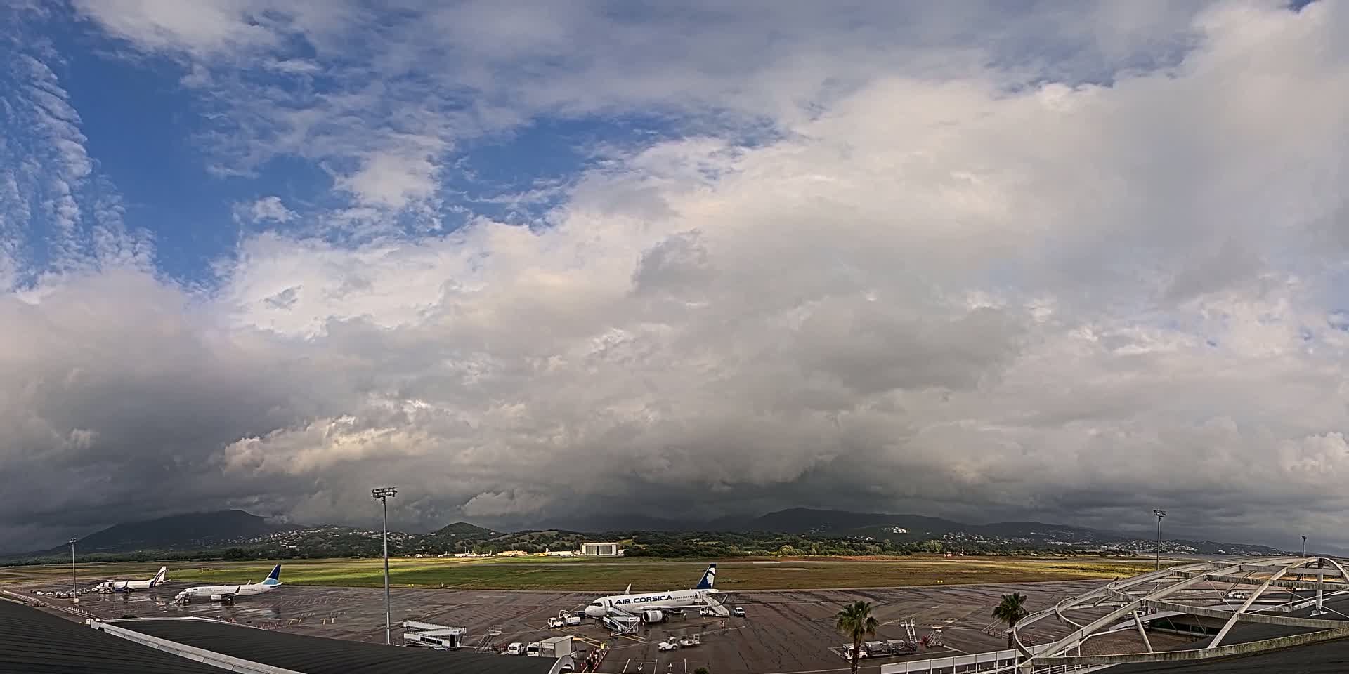 Several airplanes are parked on an airport tarmac under a mostly cloudy sky, with dark storm clouds building in the distance over mountains.