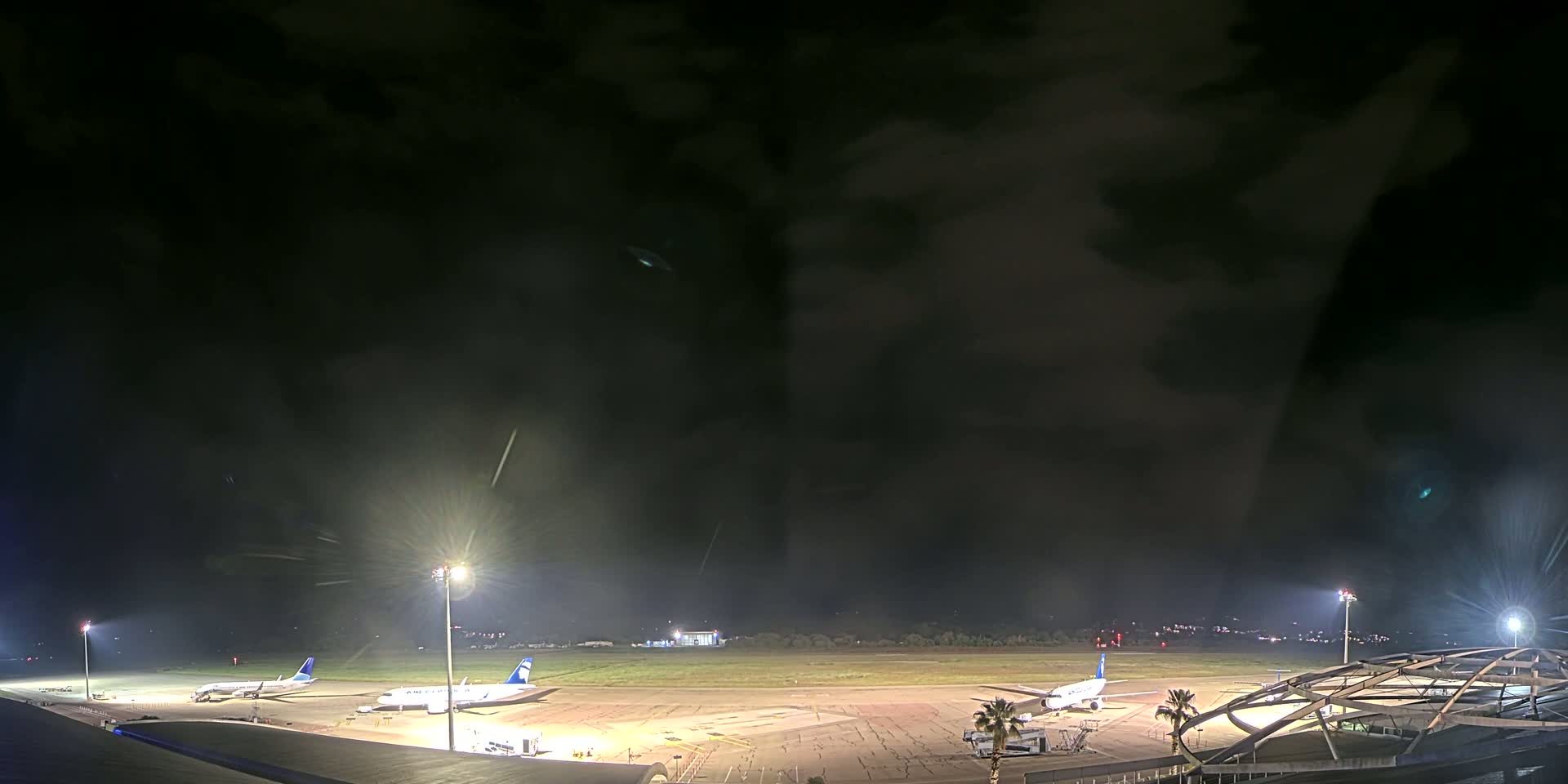 At night, under a dark, cloudy sky, several airplanes are parked on a brightly lit airport tarmac.