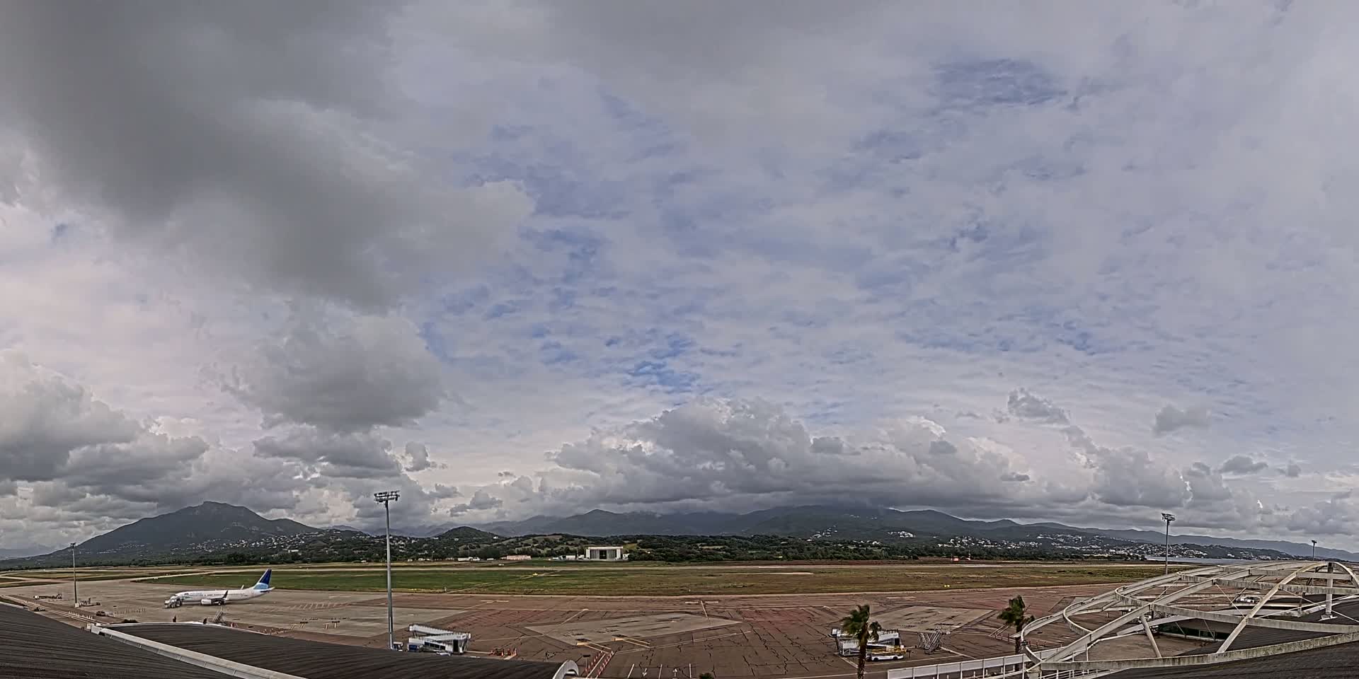 A mostly cloudy sky hangs over an airport tarmac with a single airplane parked and mountains in the distance.