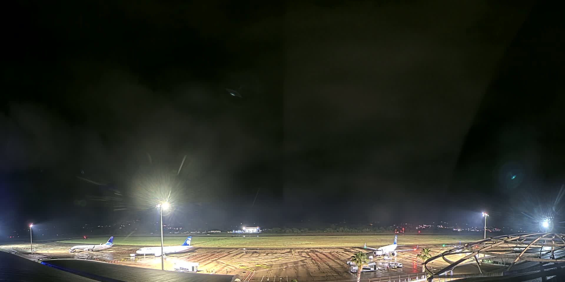 At night, under a dark, cloudy sky, several airplanes are parked on a rain-slicked airport tarmac illuminated by bright lights.