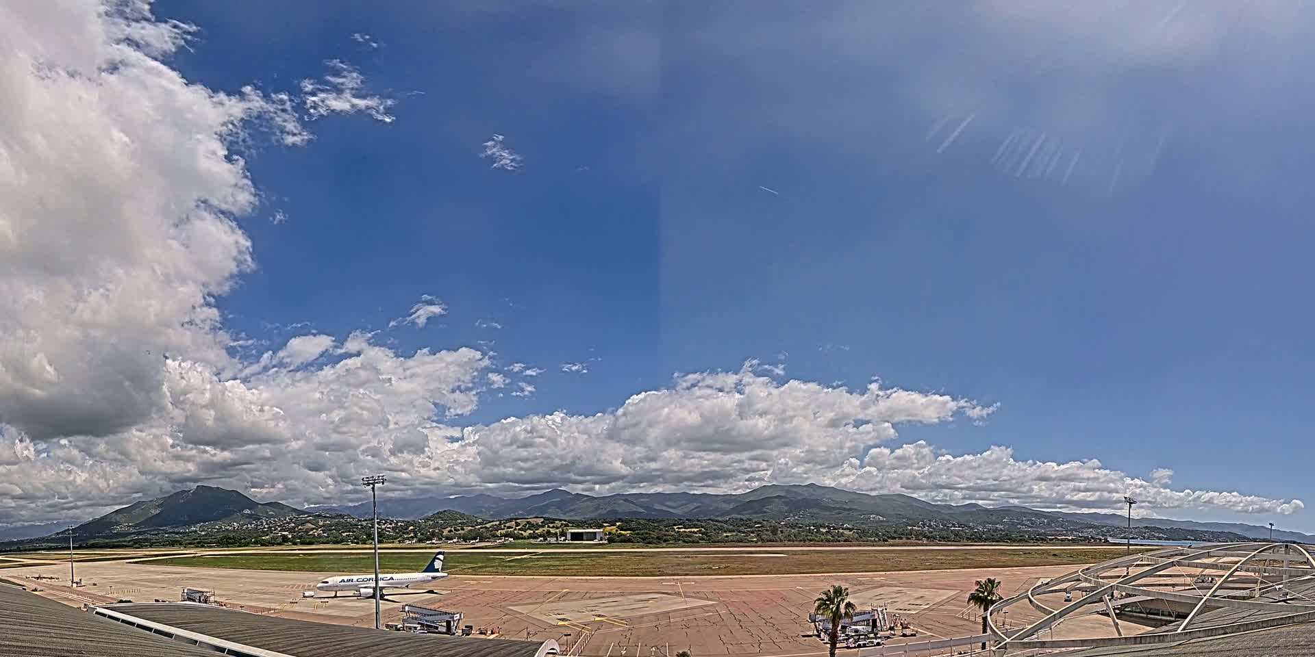 A partly cloudy sky over an airport tarmac with a passenger plane and mountains in the distance.