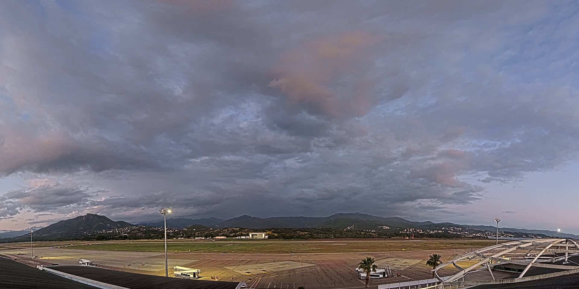 An airport tarmac and runway are visible beneath a mostly cloudy sky, with mountains in the background.